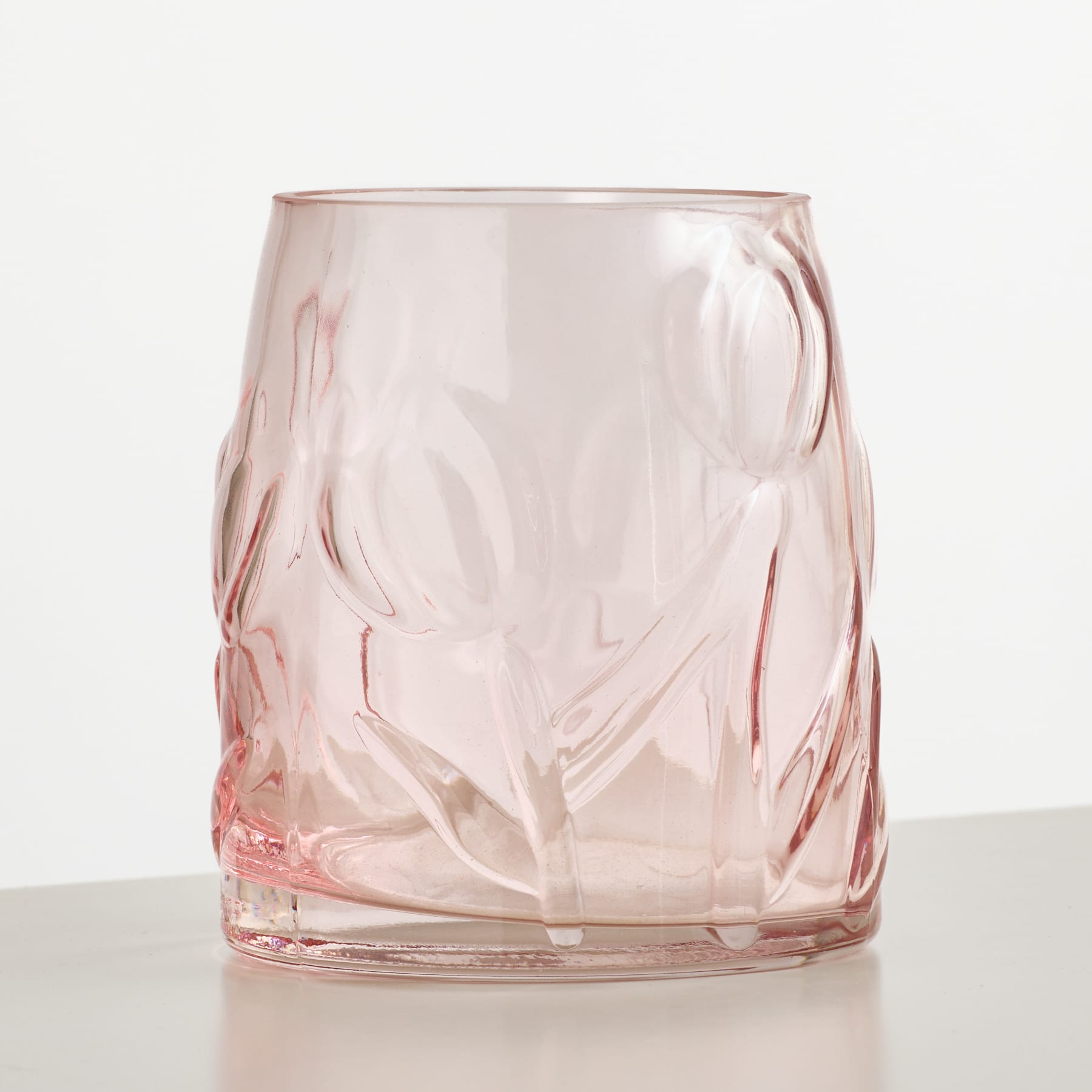 Eine rosafarbene Glasvase mit geprägten Blumenmustern steht auf einer weißen Fläche vor einem schlichten weißen Hintergrund.