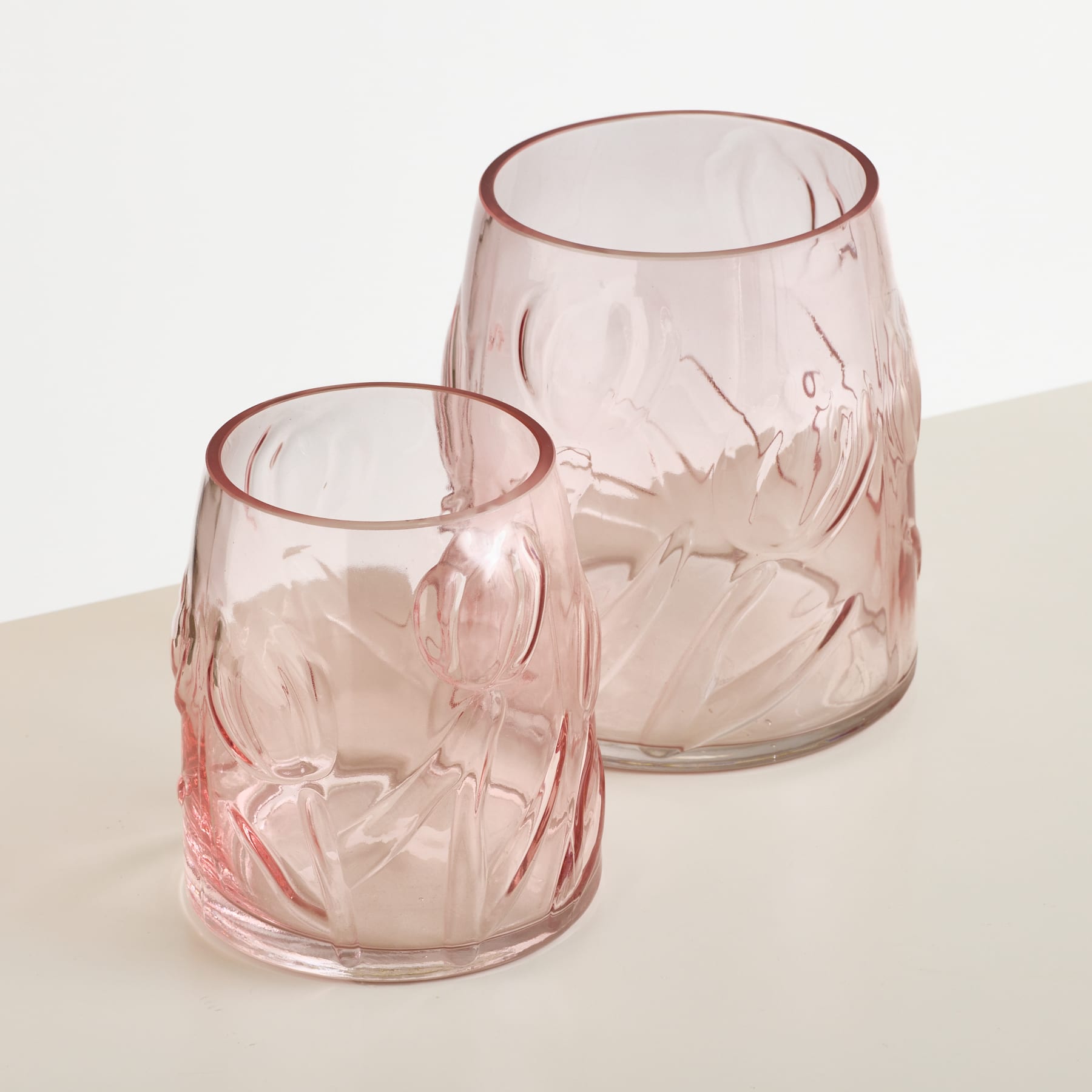 Zwei rosafarbene Glasvasen mit geprägten Blumenmustern stehen auf einer weißen Fläche vor einem schlichten weißen Hintergrund.