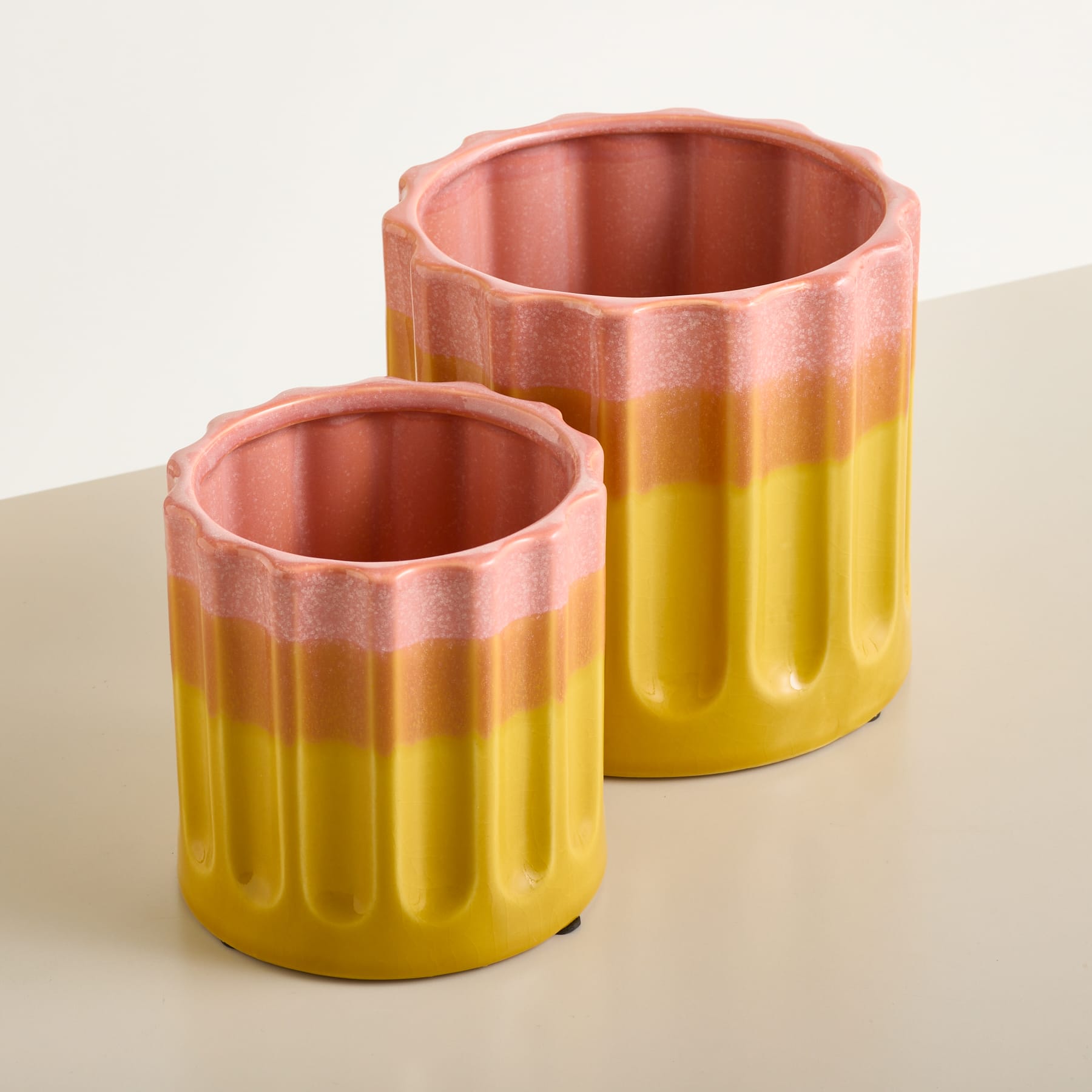 Zwei gerippte Keramiktöpfe, einer kleiner als der andere, mit rosa Rand und gelbem Korpus, stehen auf einer hellen Oberfläche vor einem sanften, neutralen Hintergrund.
