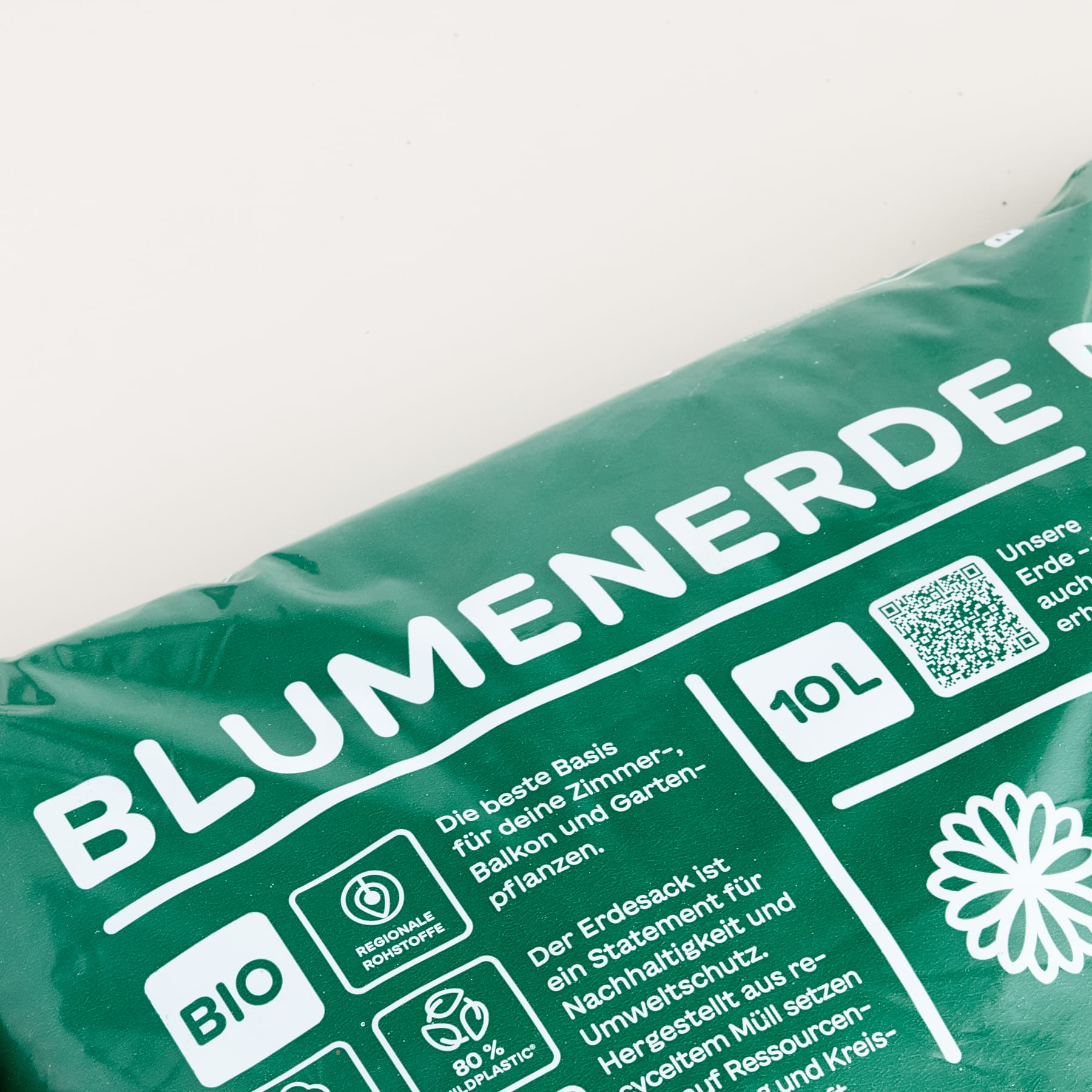 Ein grüner Sack Blumenerde mit deutscher Aufschrift ist im Vordergrund vor einem schlichten weißen Hintergrund schräg zu sehen.