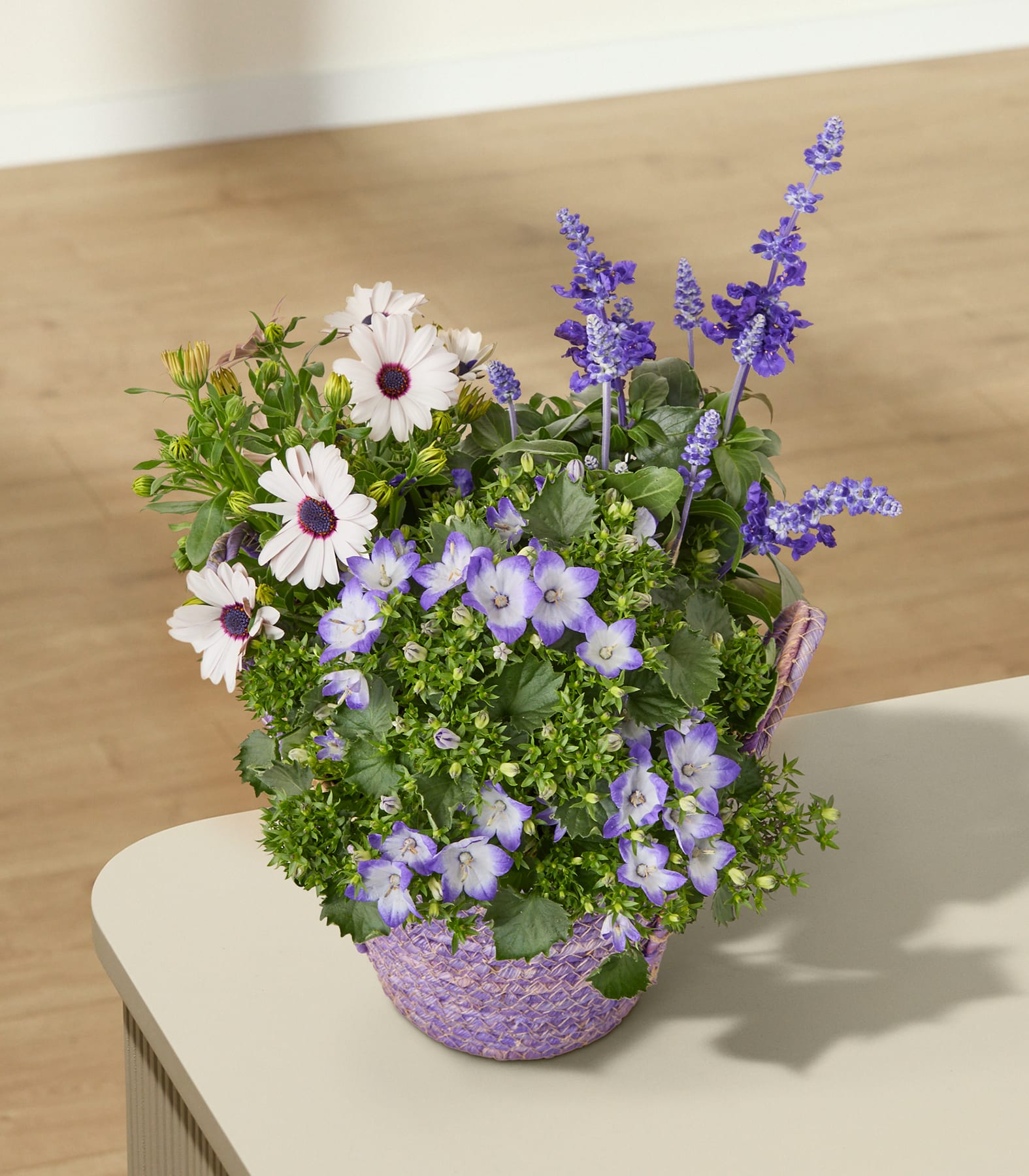 Ein farbenfroher Strauß violetter und weißer Blumen, darunter Lavendel und Gänseblümchen, arrangiert in einem geflochtenen Korb auf einer hellen Oberfläche.