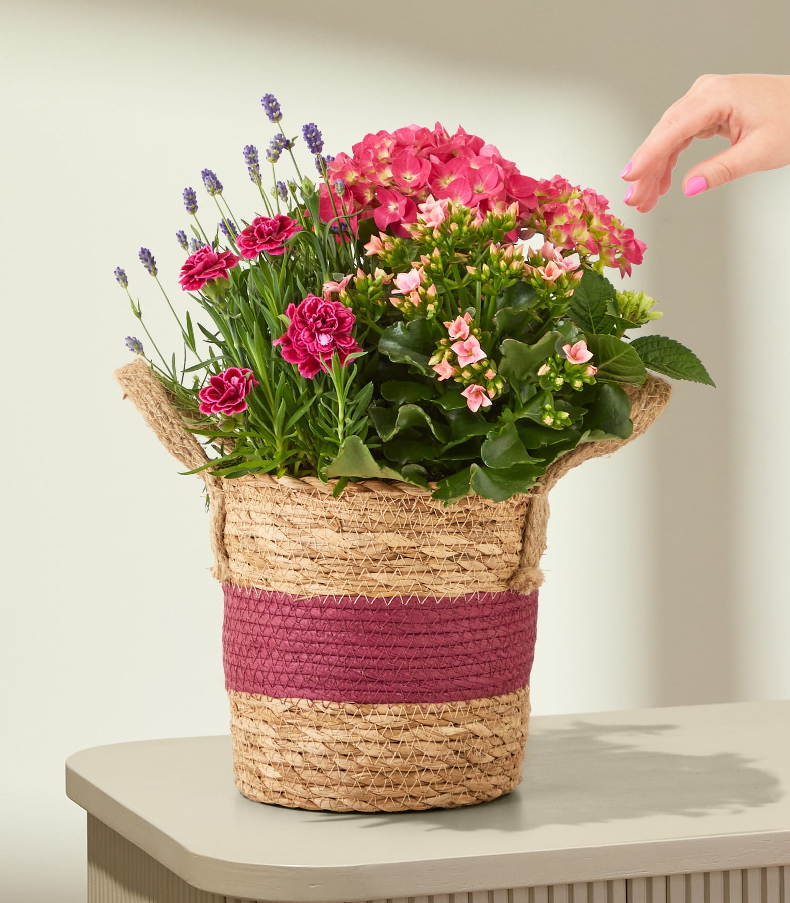 Auf einem Tisch steht ein geflochtener Korb, gefüllt mit rosa Hortensien, Nelken und Lavendel; im Hintergrund ist eine Hand zu sehen, die danach greift.