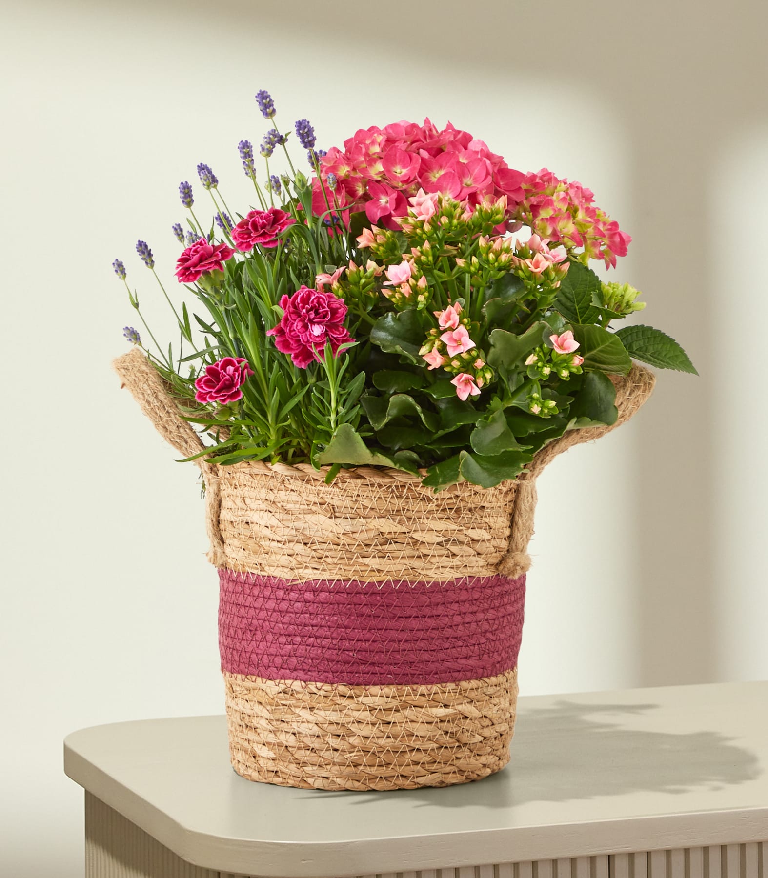 Ein geflochtener Korb, gefüllt mit rosa Hortensien, Nelken und Lavendel, steht auf einer hellen Oberfläche vor einem sanft beleuchteten, strukturierten Hintergrund.