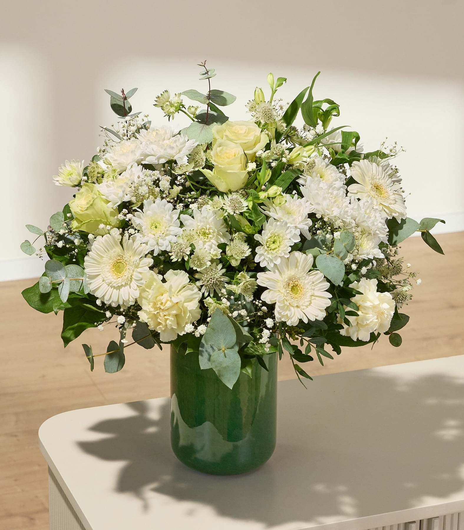 Ein lebhafter Strauß aus weißen und hellrosa Blumen, darunter Rosen, Gänseblümchen und anderes Grün, füllt das Bild vor einem schlichten weißen Hintergrund.