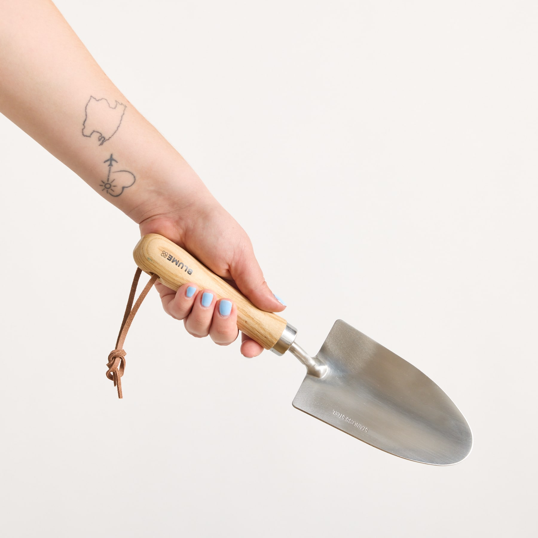 Ein tätowierter Arm hält eine Metallkelle mit Holzgriff vor einem schlichten weißen Hintergrund.
