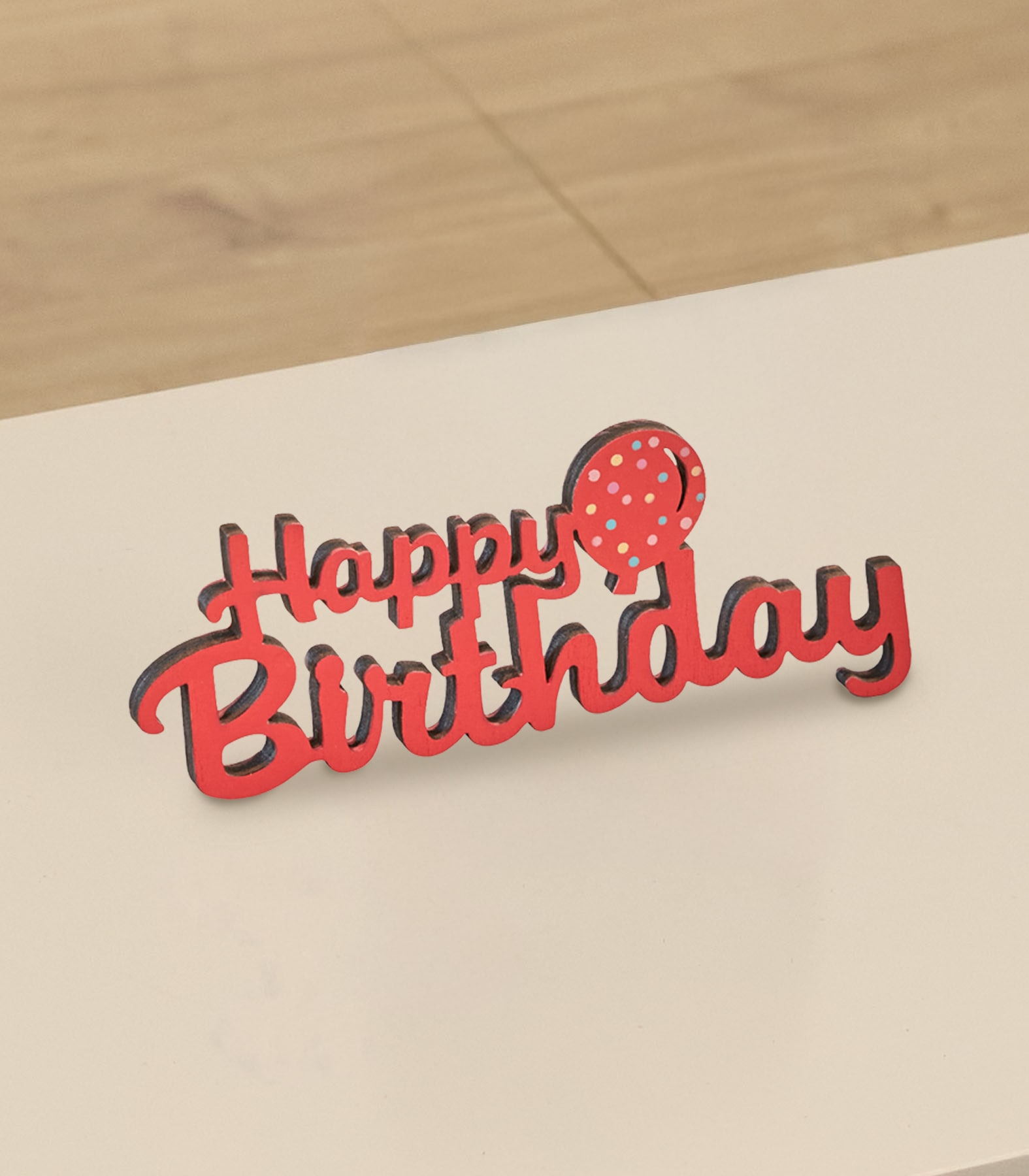 Ein roter „Happy Birthday“-Kuchenaufsatz mit einem gepunkteten Ballon steht auf einer hellen Oberfläche vor einem verschwommenen Holzhintergrund.