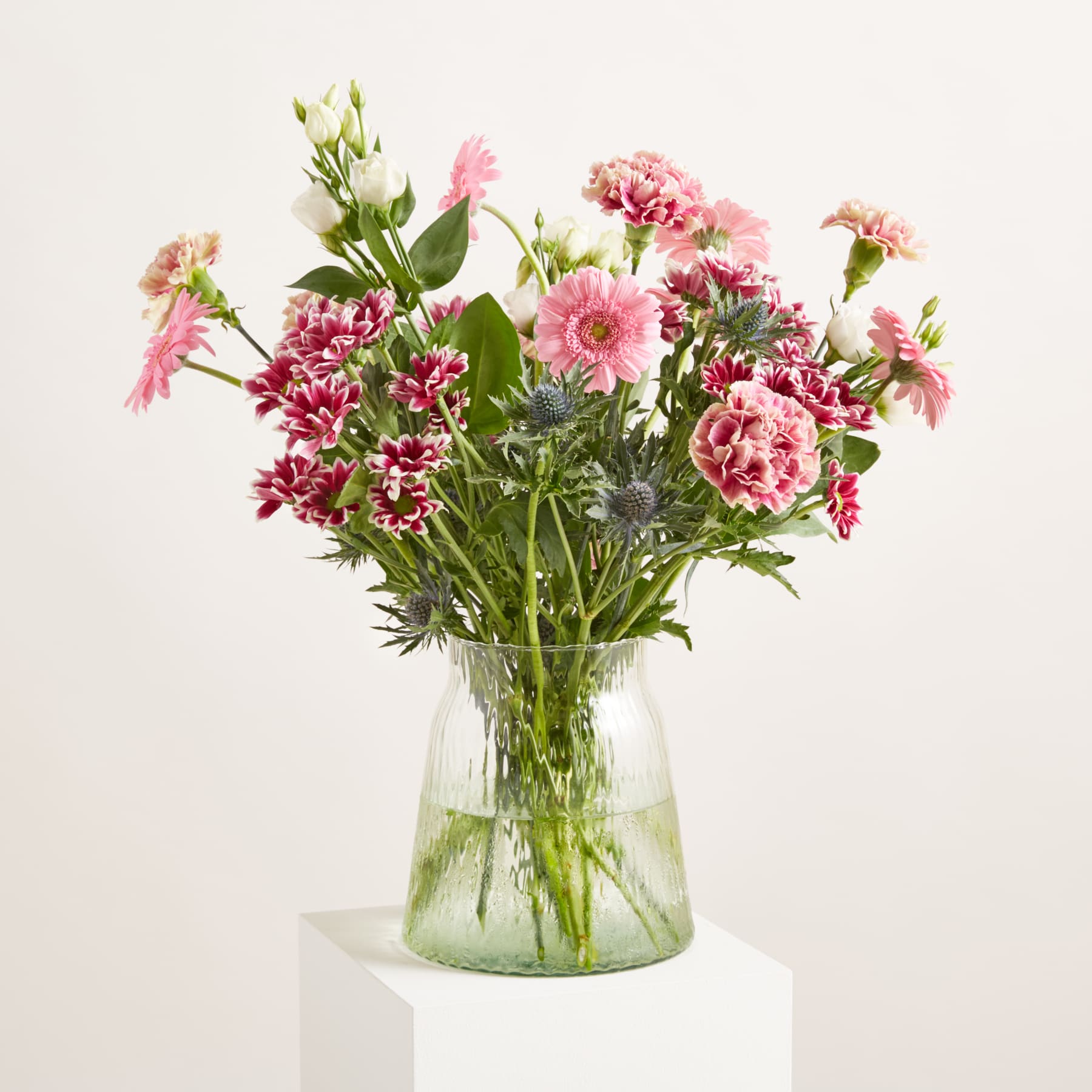 Ein farbenfroher Strauß aus rosa und weißen Blumen, darunter Gerbera und Nelken, ist in einer durchsichtigen, gerippten Glasvase vor einem schlichten weißen Hintergrund arrangiert.