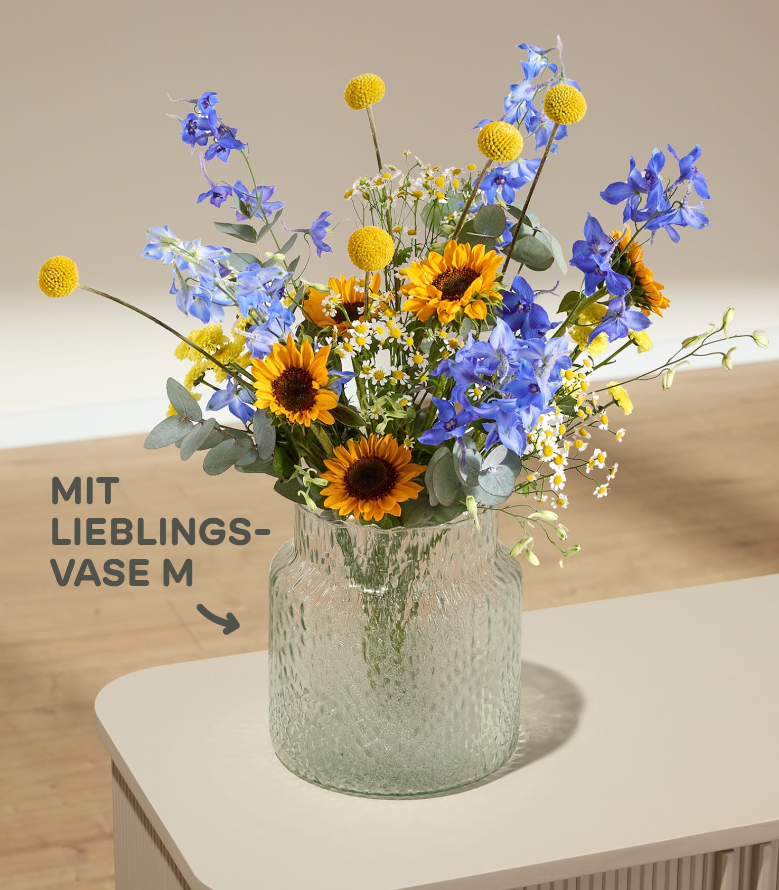 Im Vordergrund steht eine strukturierte Glasvase mit einem farbenfrohen Strauß aus Sonnenblumen, blauen Ritterspornen und gelben Knopfblumen vor einem sanft beleuchteten, neutralen Hintergrund mit deutschem Text.