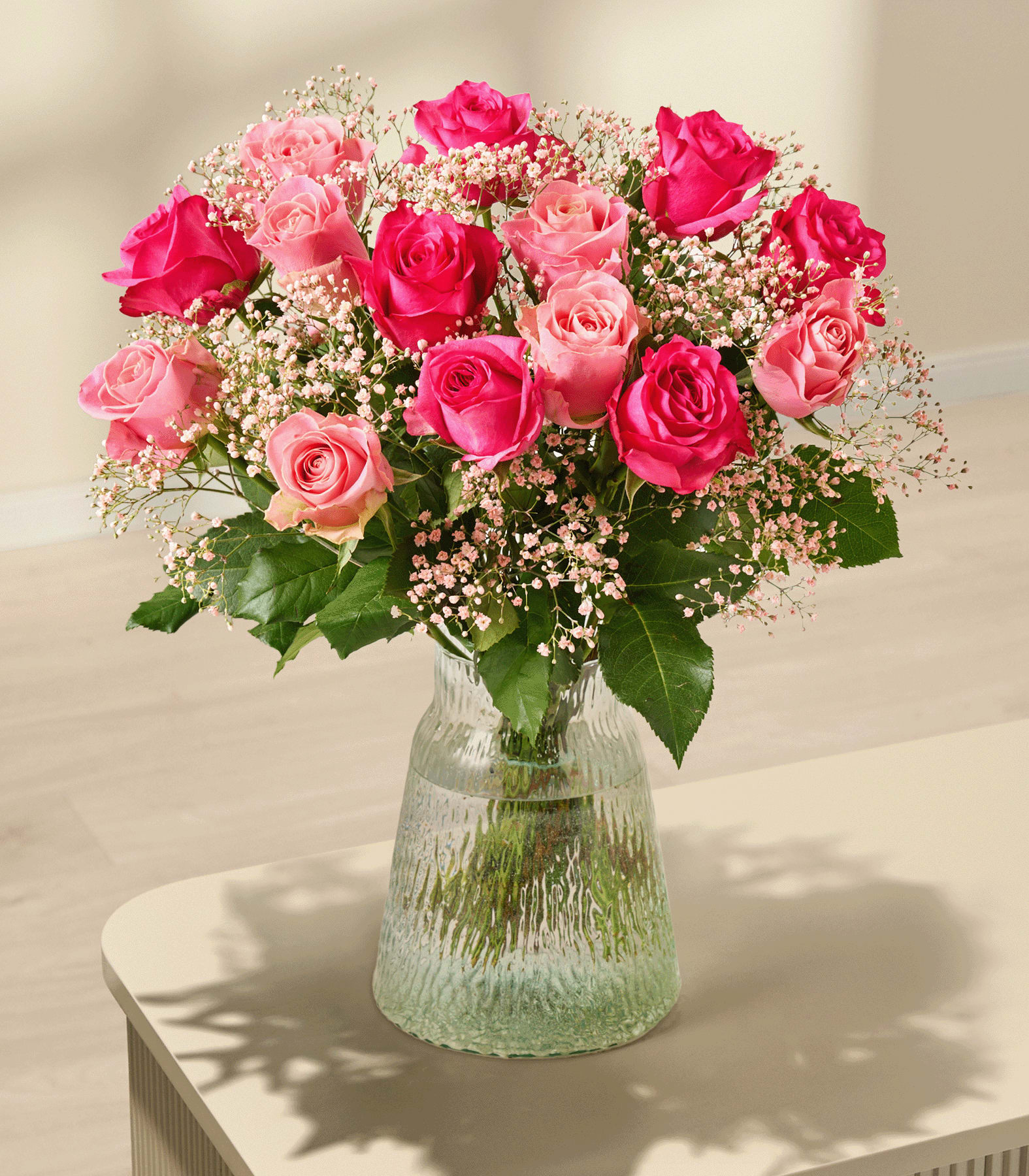 Ein Strauß aus rosa und roten Rosen mit Schleierkraut füllt eine strukturierte Glasvase auf einem hellen Tisch vor einem sanft beleuchteten, neutralen Hintergrund.