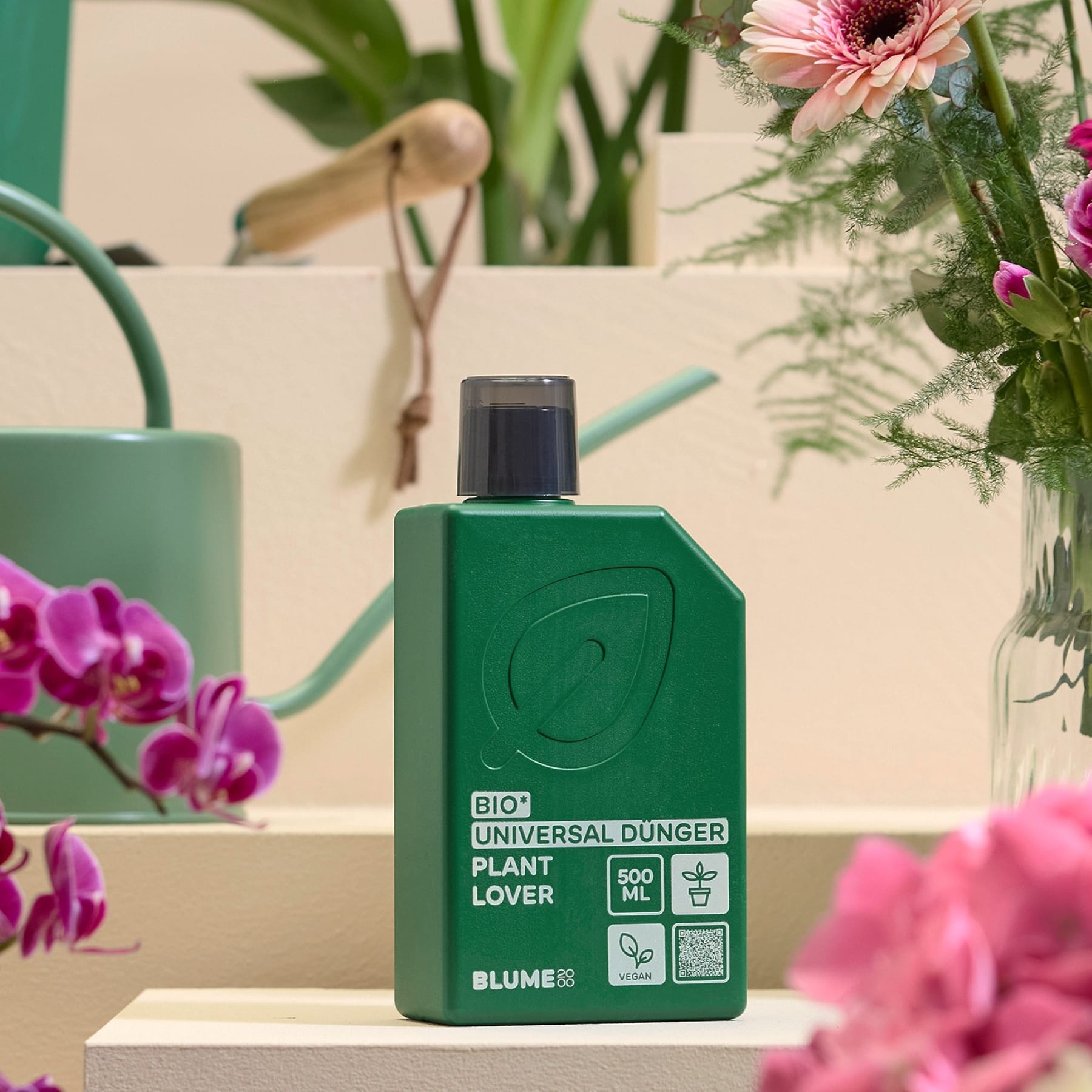 Auf einer cremefarbenen Stufe steht eine grüne Flasche Pflanzennahrung, umgeben von leuchtend pinkfarbenen Orchideen und Hortensien; im verschwommenen Hintergrund sind eine Gießkanne und Gartengeräte zu sehen.