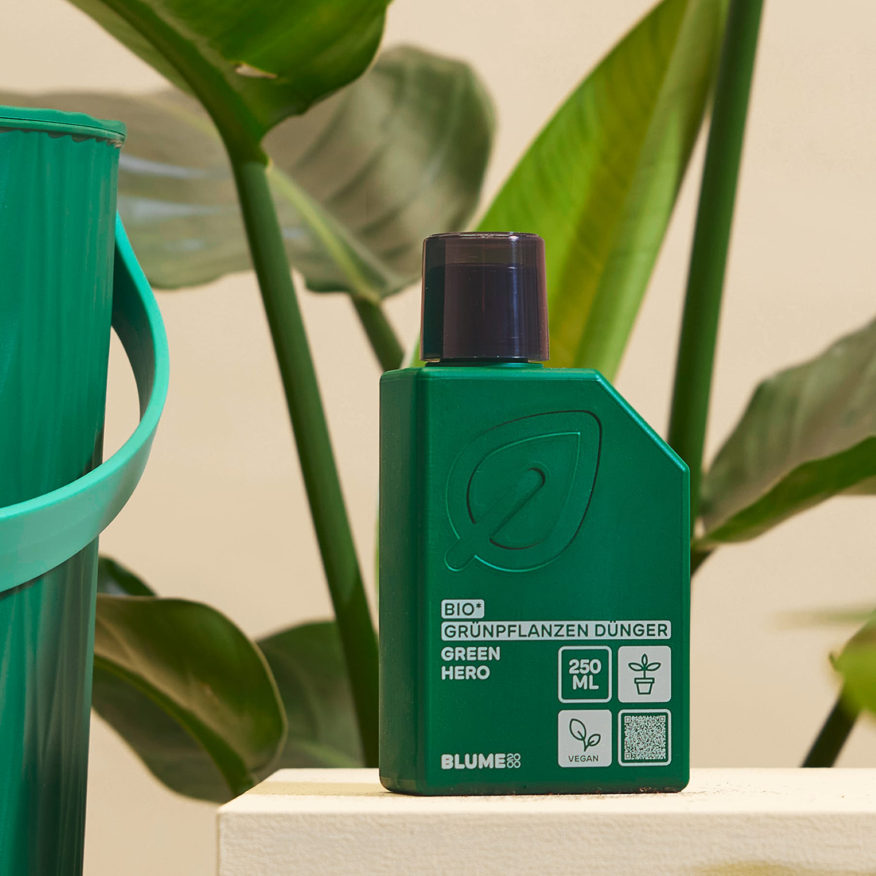 Im Vordergrund steht eine grüne Flasche Pflanzendünger auf einer hellen Oberfläche, im Hintergrund sind ein verschwommener grüner Eimer und große Pflanzenblätter zu sehen.