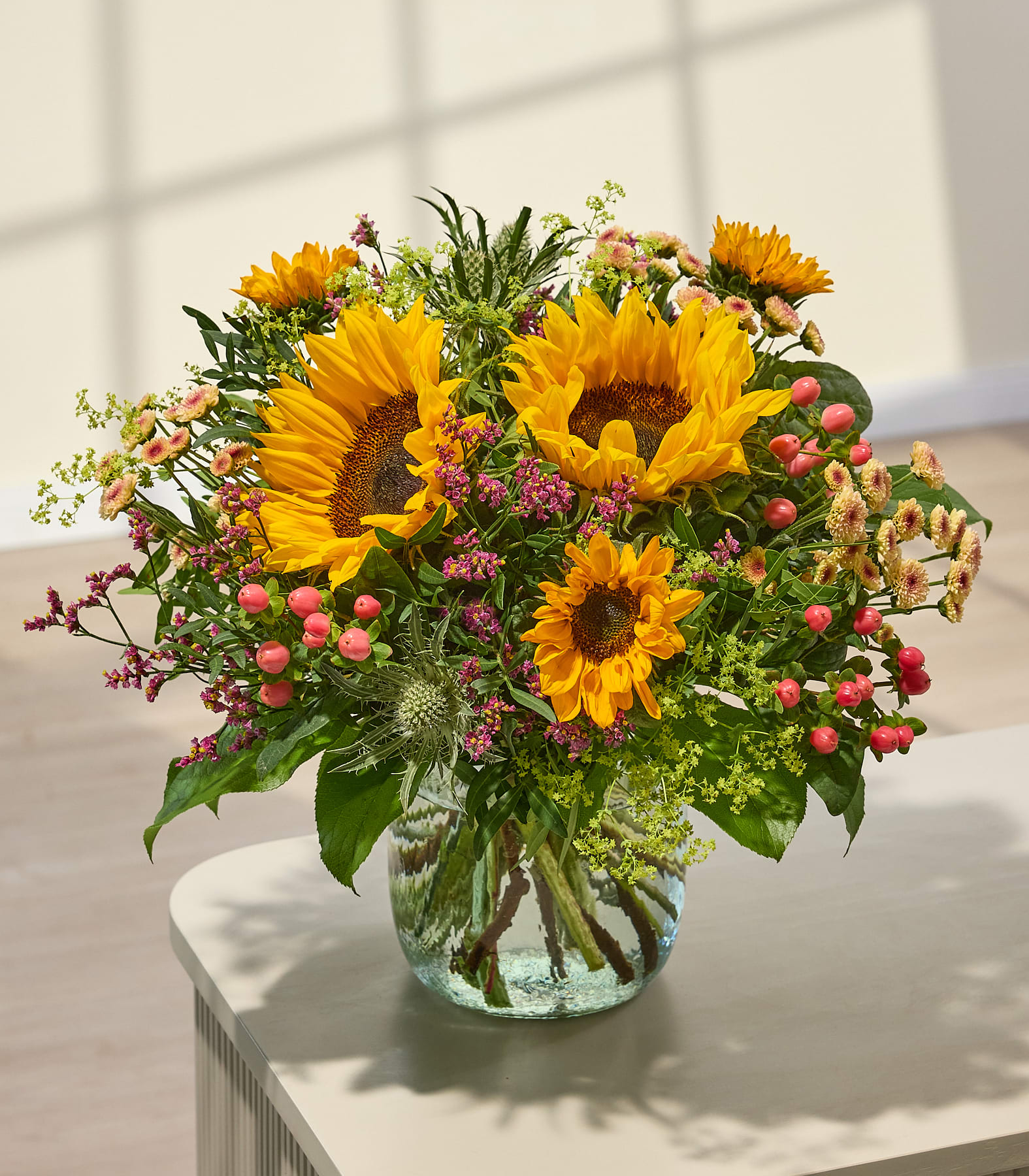 Ein leuchtender Strauß Sonnenblumen und Wildblumen steht in einer Glasvase auf einem Tisch, während im Hintergrund sanftes Licht und Schatten eine warme Atmosphäre schaffen. (Beschreibungstext KI-generiert)