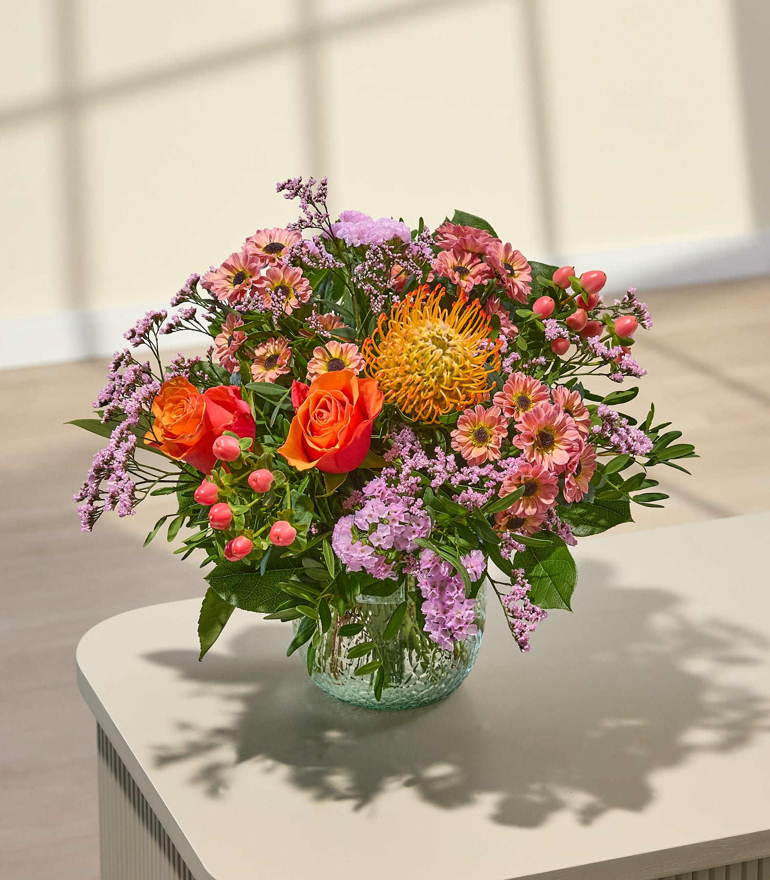 Ein leuchtender Strauß orangefarbener und rosafarbener Blumen steht in einer strukturierten Glasvase auf einem hellen Tisch, im Hintergrund werfen sanfte Schatten sanfte Schatten auf eine helle Wand. (Beschreibungstext KI-generiert)