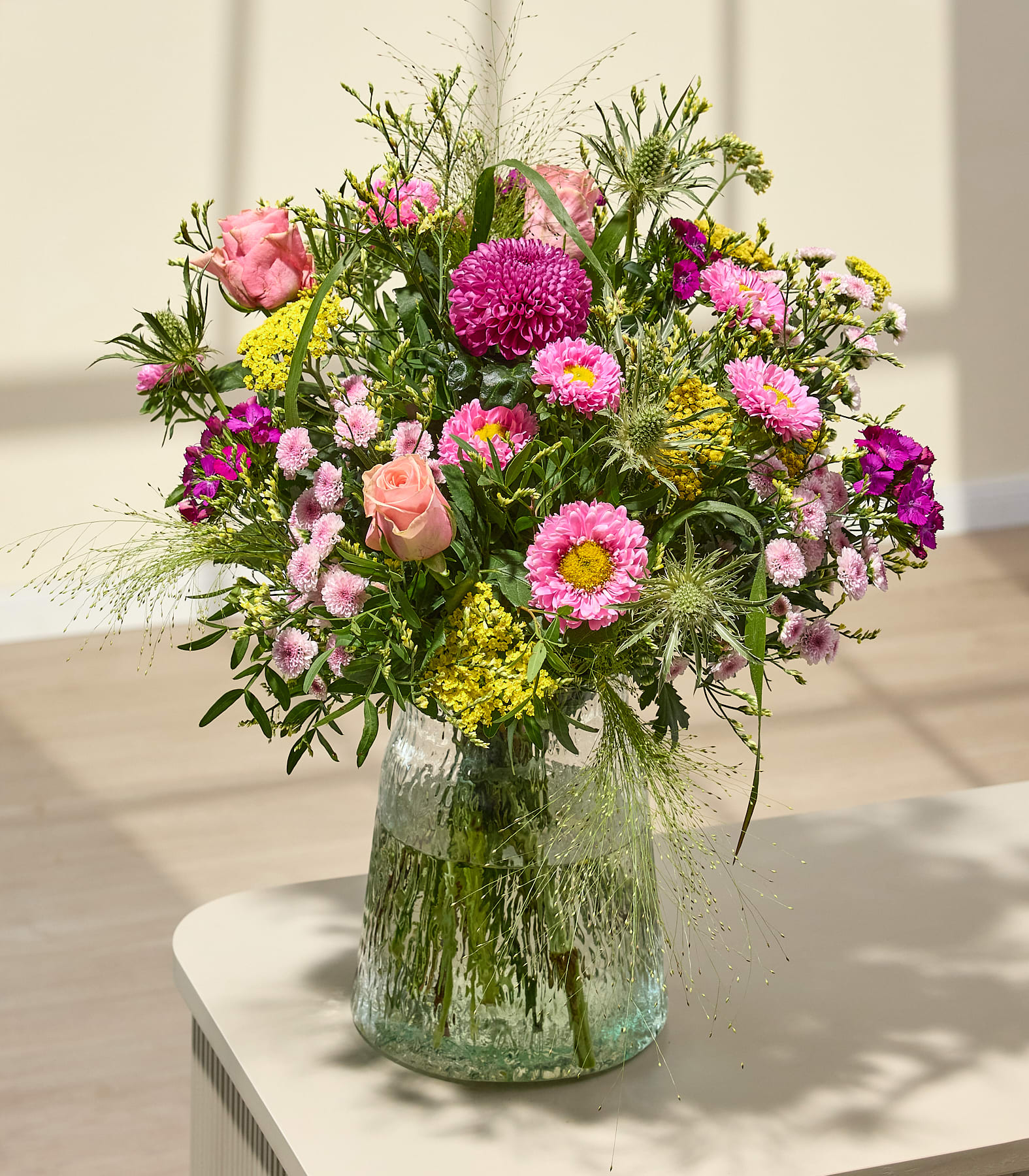 Im Vordergrund füllt ein leuchtender Strauß rosa und gelber Blumen eine strukturierte Glasvase vor einem sanft beleuchteten, neutralen Hintergrund. (Beschreibungstext KI-generiert)
