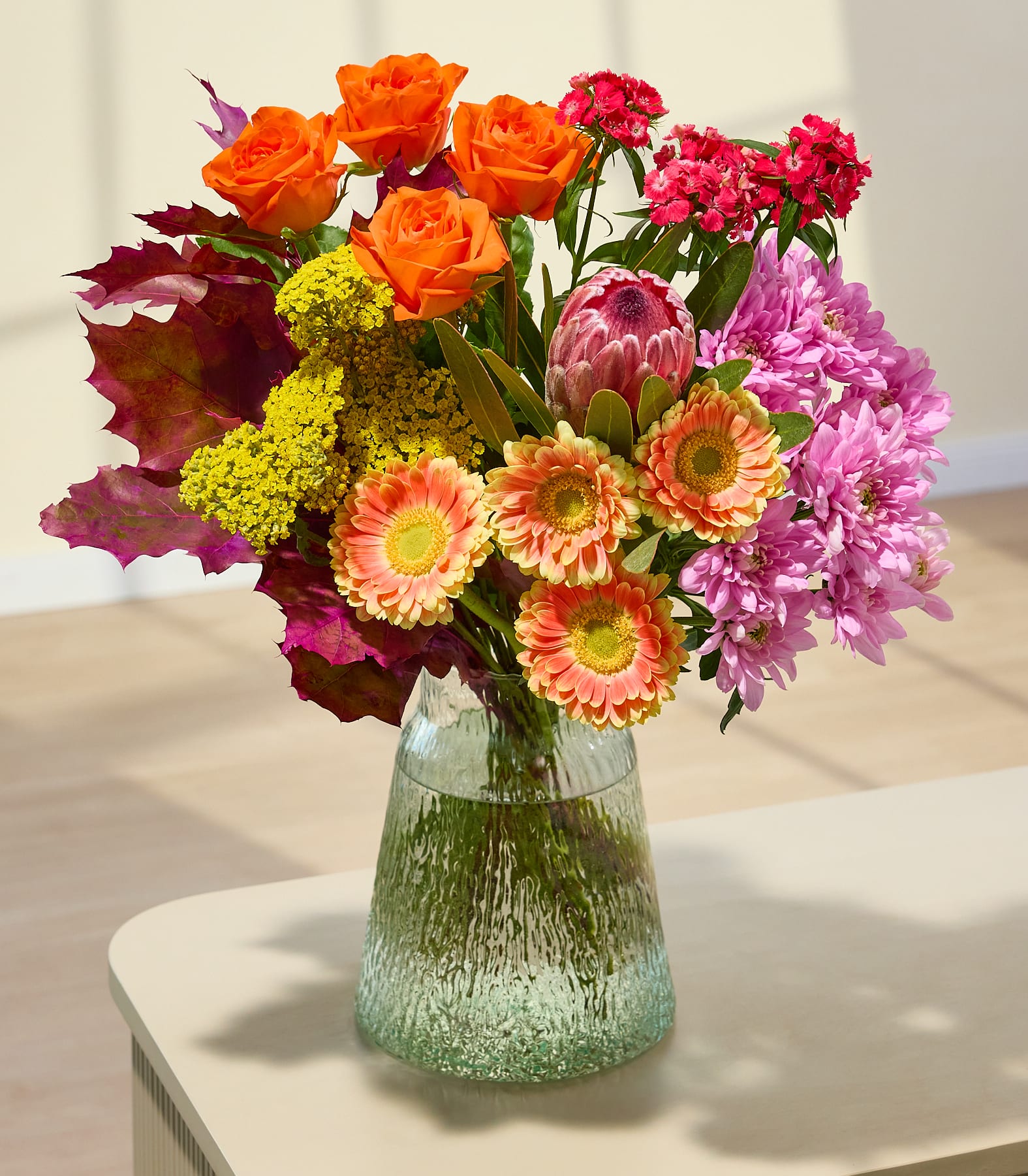 Ein farbenfroher Strauß aus verschiedenen rosa und orangefarbenen Blumen, darunter Rosen, Nelken und Chrysanthemen, der von einer Person in den Händen gehalten wird, vor einem schlichten weißen Hintergrund. (Beschreibungstext KI-generiert)