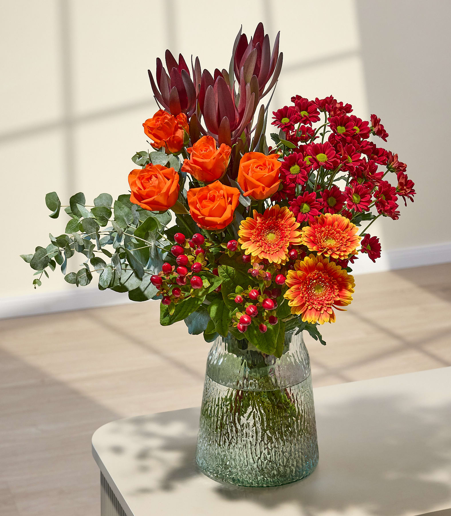 Ein farbenfroher Blumenstrauß aus verschiedenen Blumen, darunter rote Gerbera-Gänseblümchen, orangefarbene Rosen und violette Proteen, in den Händen einer Person vor einem schlichten weißen Hintergrund.