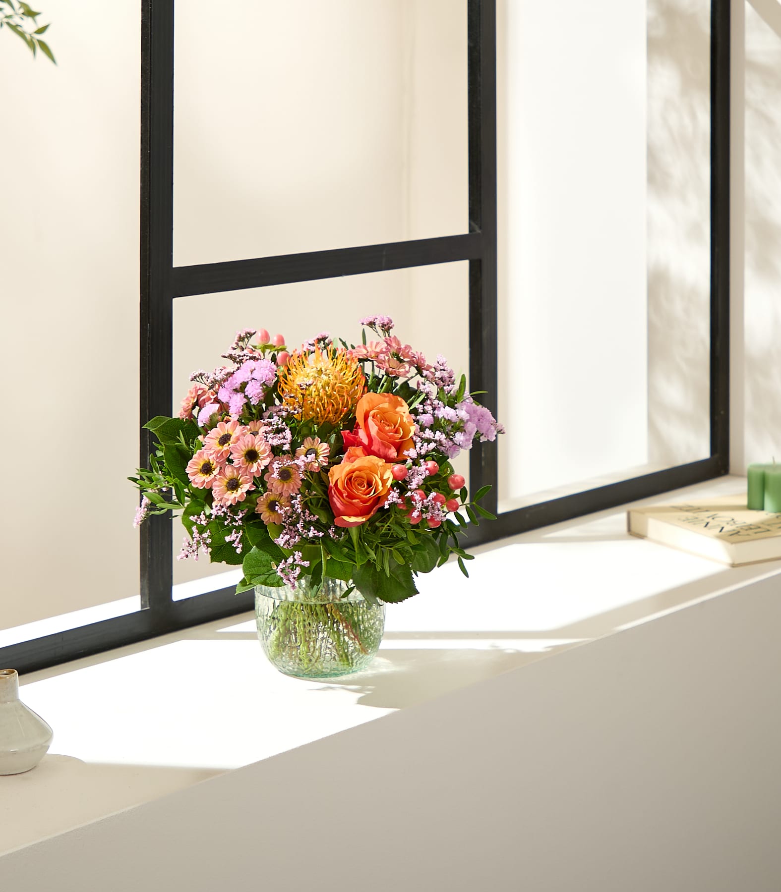 Ein leuchtender Strauß orangefarbener und rosafarbener Blumen steht in einer Glasvase auf einer Fensterbank, im Hintergrund strömt Sonnenlicht durch ein Fenster. (Beschreibungstext KI-generiert)