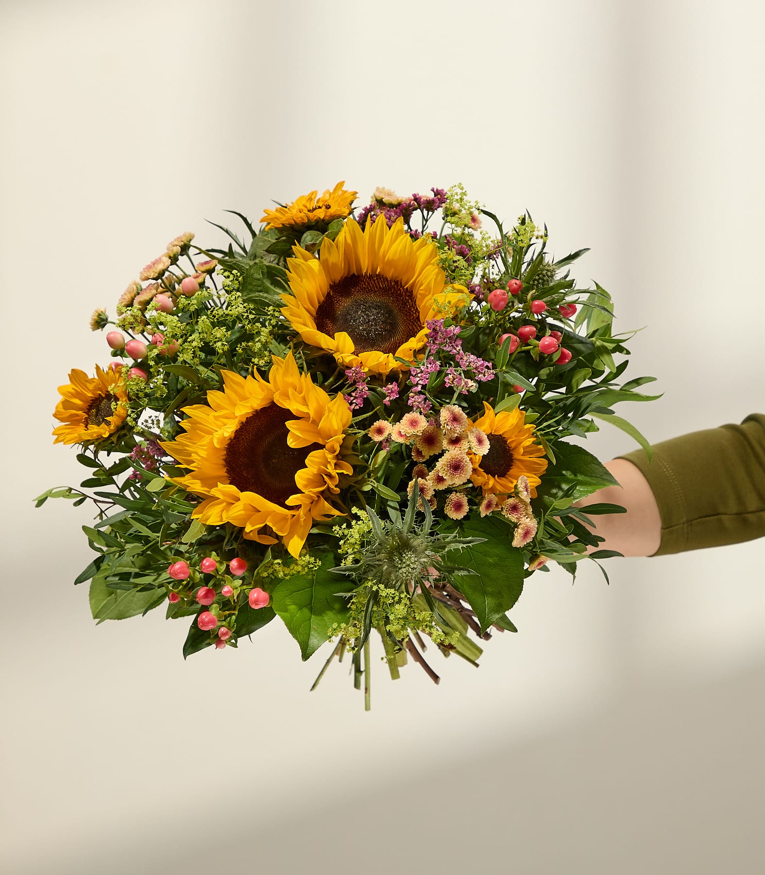 Eine Hand hält einen leuchtenden Strauß Sonnenblumen und kleineren rosa und gelben Blüten vor einem sanft beleuchteten, neutralen Hintergrund. (Beschreibungstext KI-generiert)