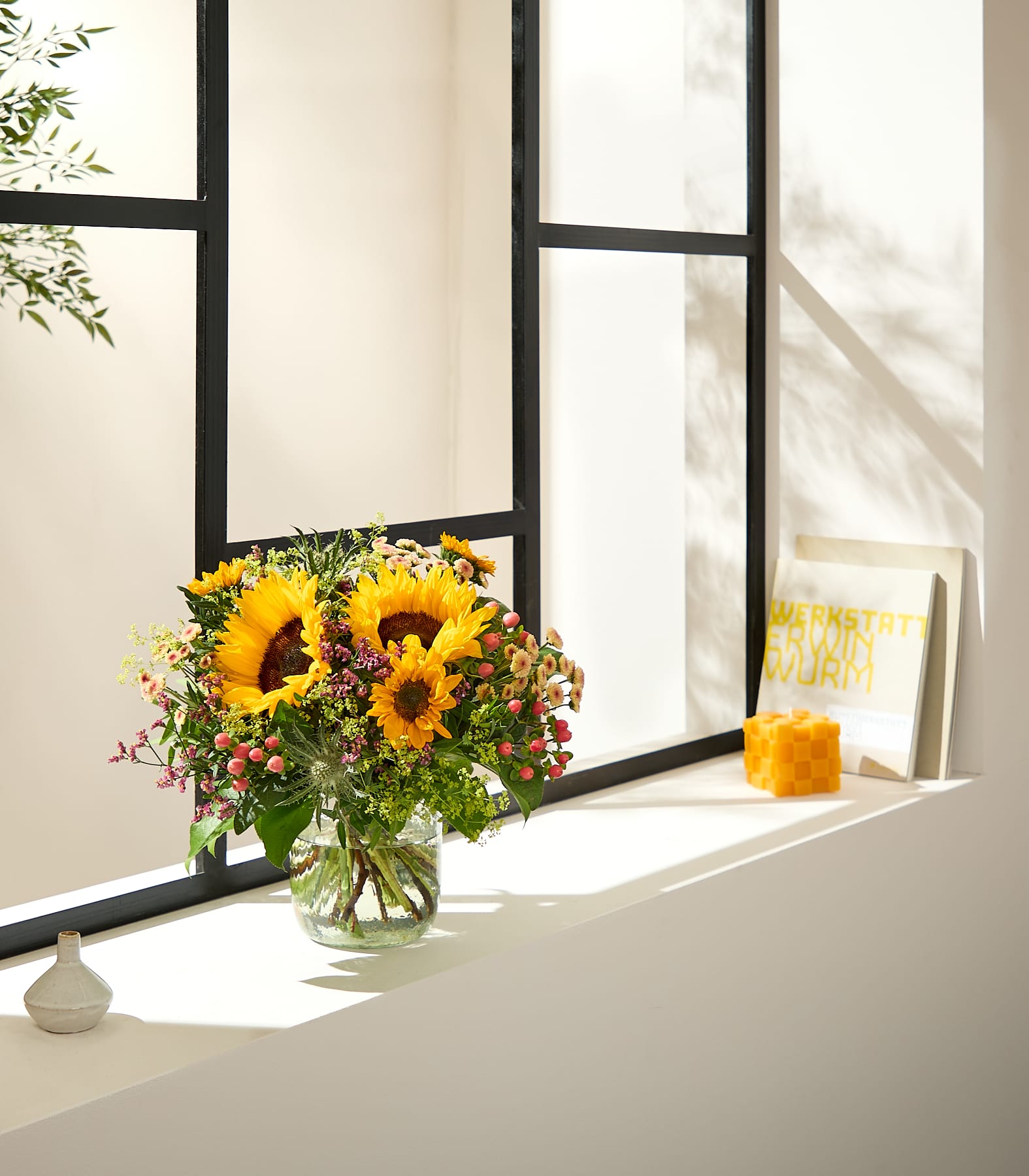 Ein leuchtender Strauß Sonnenblumen und Grünzeug steht in einer Glasvase auf dem Fensterbrett, rechts daneben gerahmte Bücher und ein kleines Deko-Objekt, alles in sanftes Sonnenlicht getaucht, das durch ein Fenster mit dunklen Sprossen fällt. (Beschreibungstext KI-generiert)