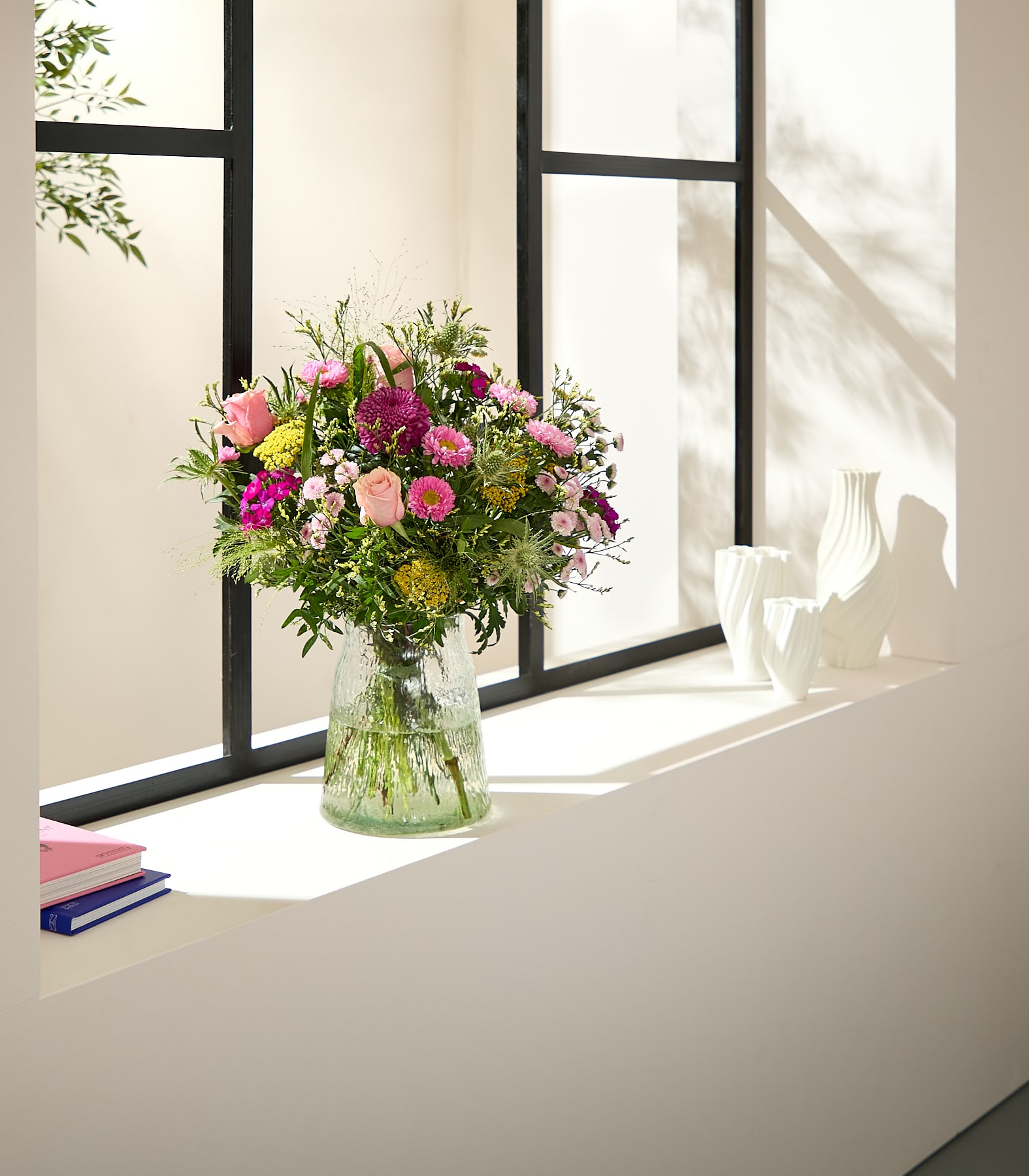 Auf einem Fensterbrett vor einem Fenster mit schwarzen Scheiben steht ein leuchtender Strauß rosa und gelber Blumen in einer Glasvase; links davon befinden sich drei weiße Vasen und zwei Bücher. (Beschreibungstext KI-generiert)