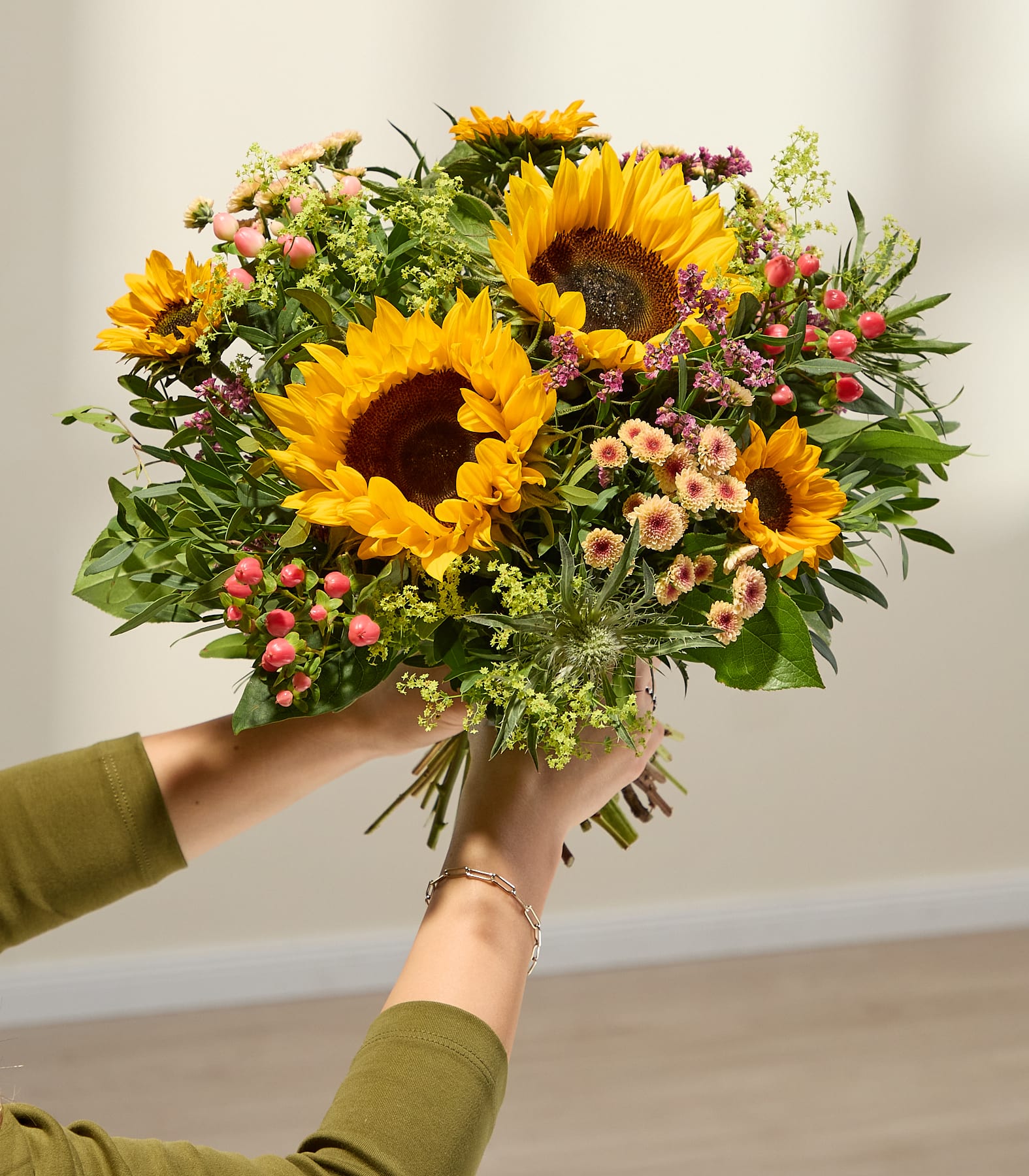 Im Vordergrund hält ein leuchtender Strauß Sonnenblumen und anderer Wildblumen die Hände vor einem sanft beleuchteten, neutralen Hintergrund. (Beschreibungstext KI-generiert)