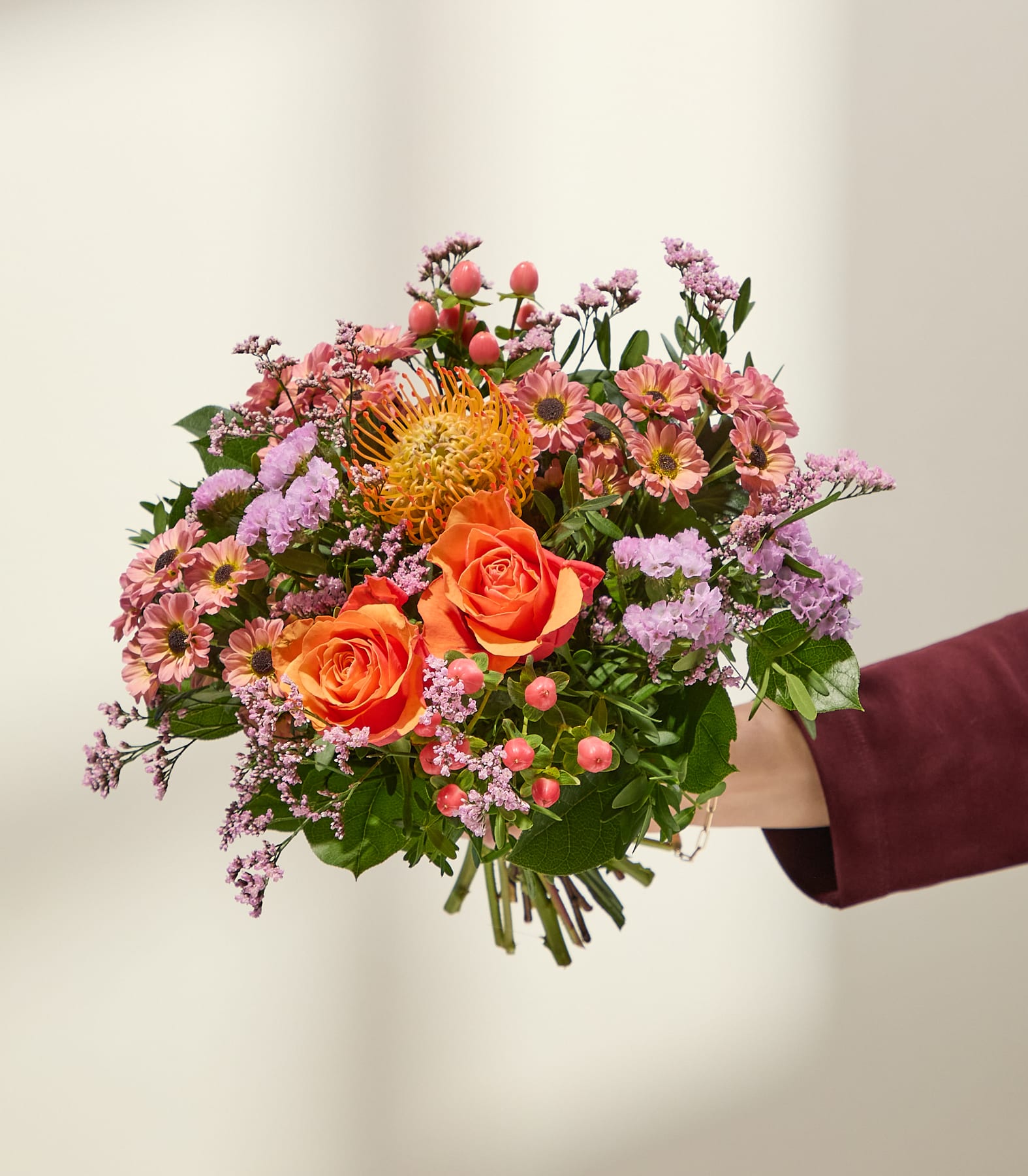 Eine Hand in einem bordeauxroten Ärmel hält einen leuchtenden Strauß aus orangefarbenen Rosen, rosa Chrysanthemen und violetten Füllblumen vor einem sanft beleuchteten, neutralen Hintergrund. (Beschreibungstext KI-generiert)