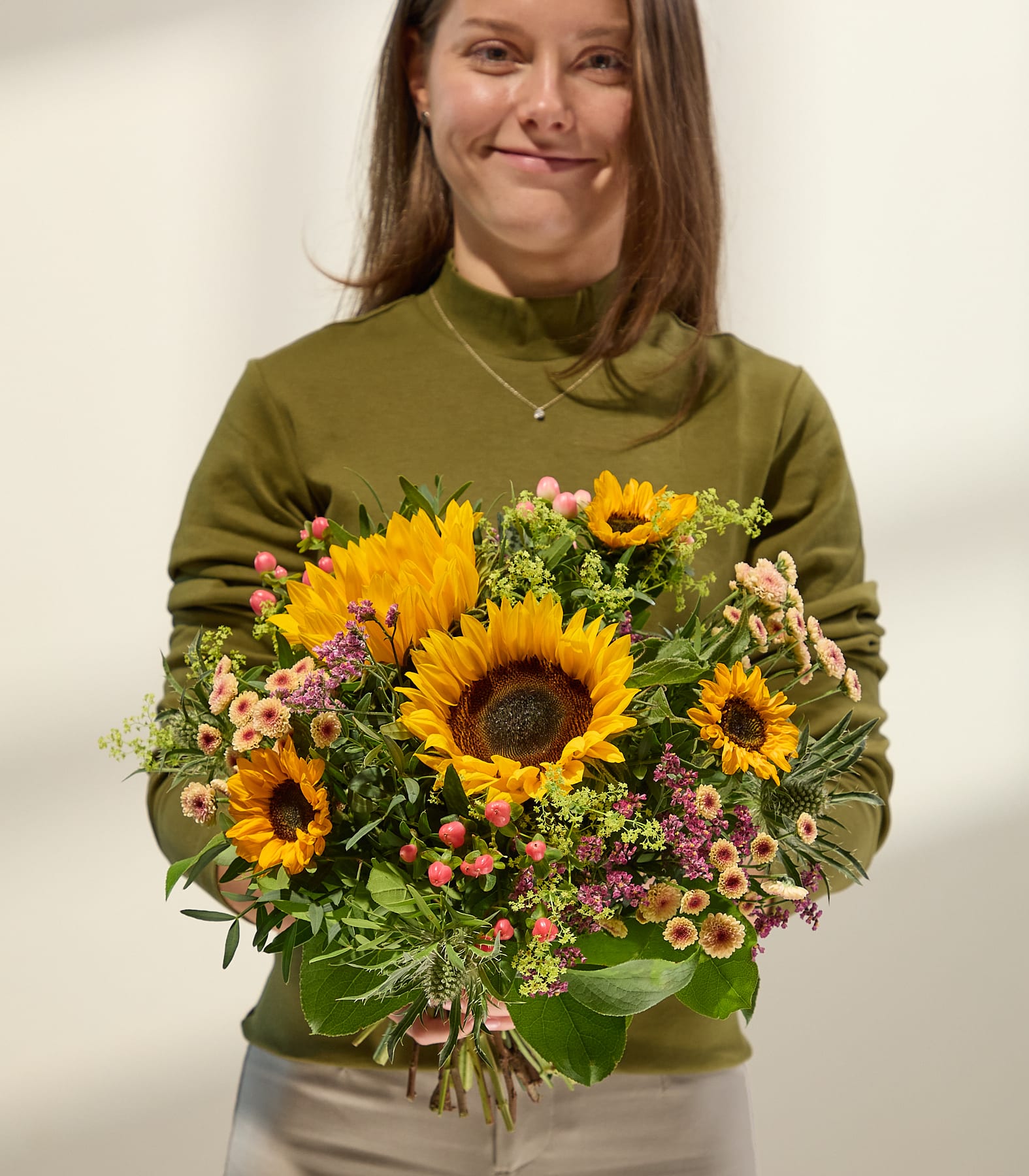 Eine lächelnde Frau in einem grünen Rollkragenpullover hält einen farbenfrohen Strauß Sonnenblumen und anderer Blumen vor einem schlichten weißen Hintergrund. (Beschreibungstext KI-generiert)