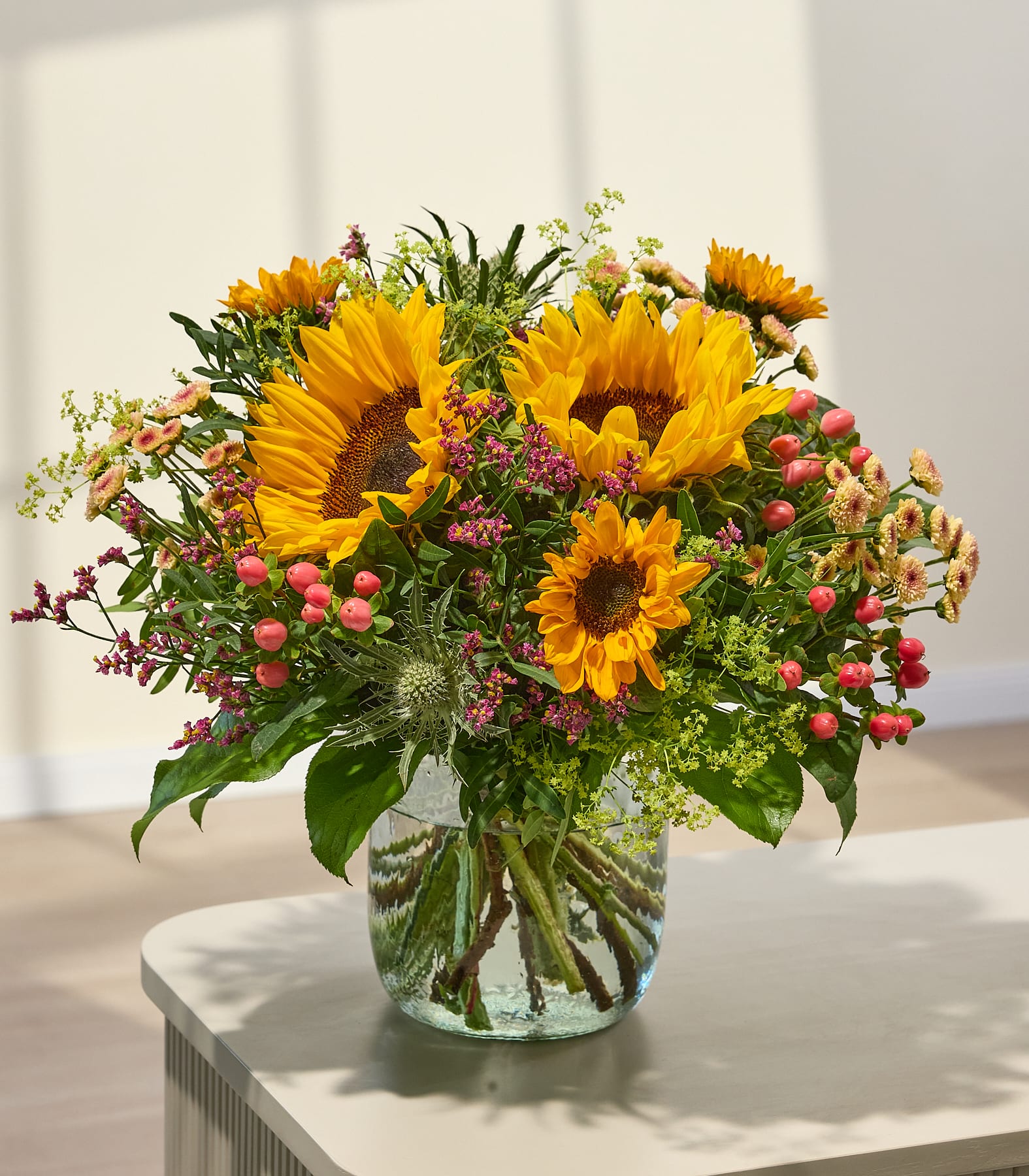 Ein farbenfroher Strauß aus Sonnenblumen und Beeren steht in einer Glasvase auf einem Tisch, im Hintergrund ist eine sanft beleuchtete Wand zu sehen. (Beschreibungstext KI-generiert)