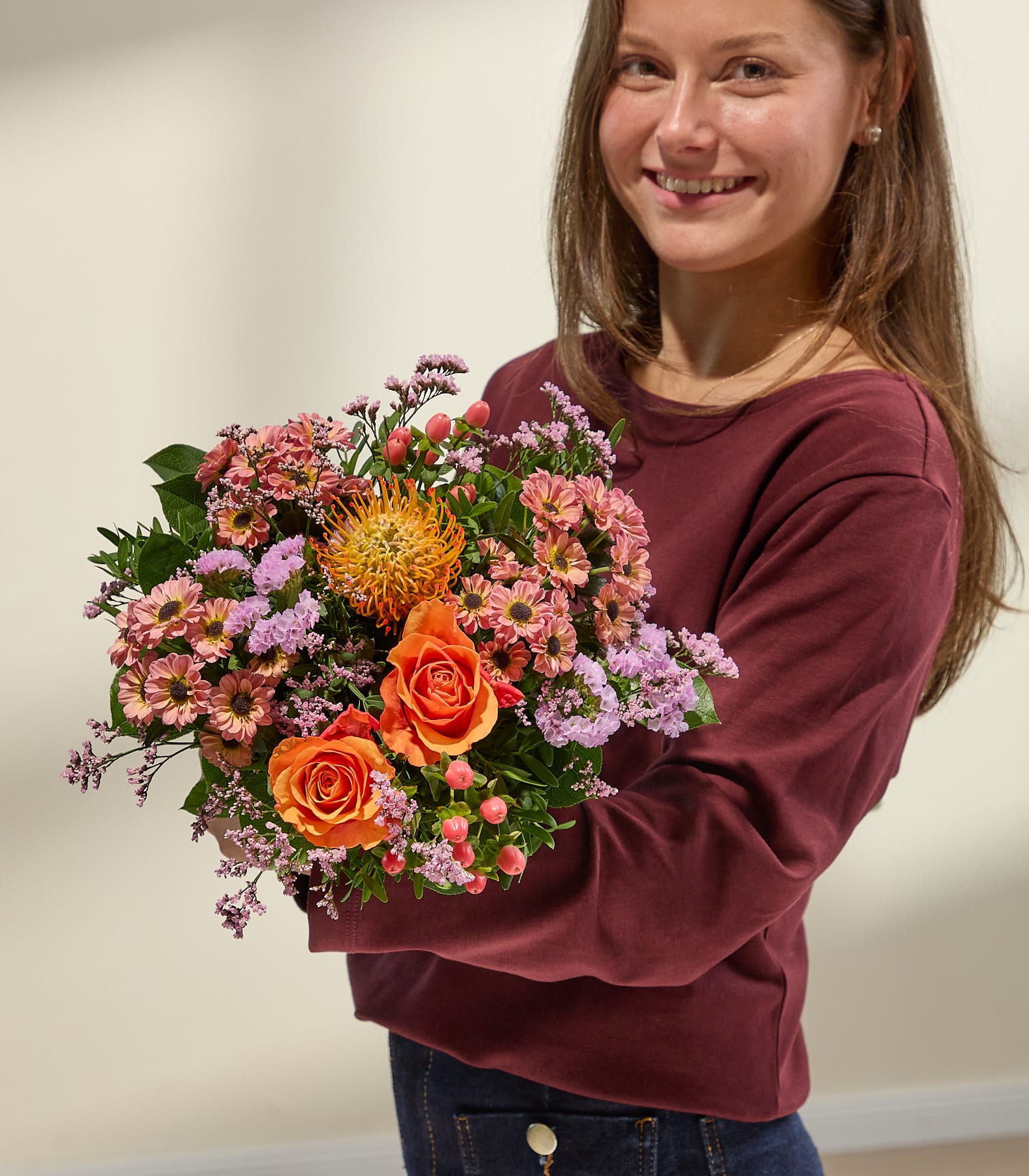 Eine lächelnde junge Frau in einem weinroten Oberteil hält vor einem sanft beleuchteten, neutralen Hintergrund einen leuchtenden Strauß orangefarbener und rosafarbener Blumen. (Beschreibungstext KI-generiert)