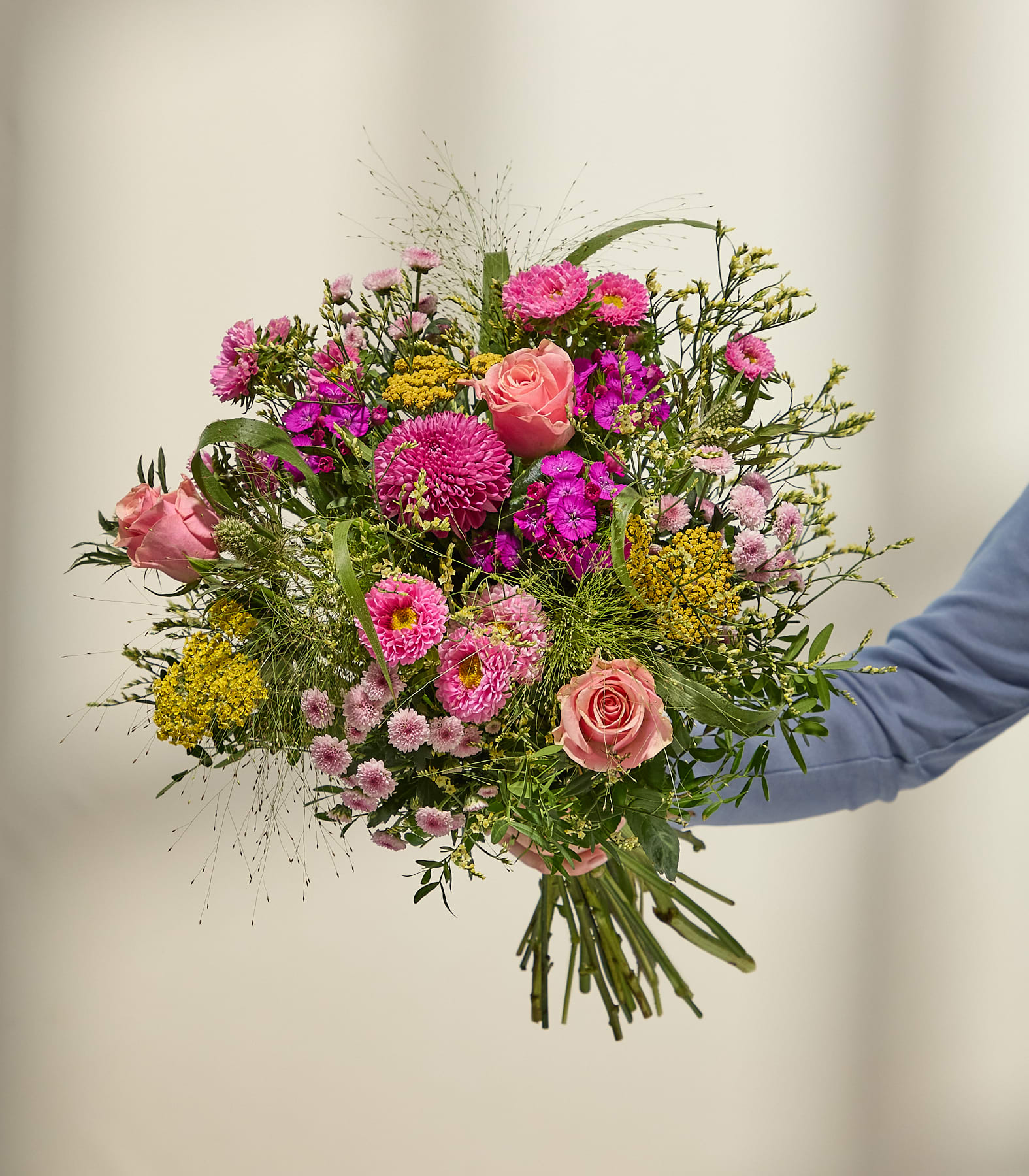 Eine Person hält einen farbenfrohen Strauß rosa und gelber Blumen vor einem sanft beleuchteten, neutralen Hintergrund. (Beschreibungstext KI-generiert)