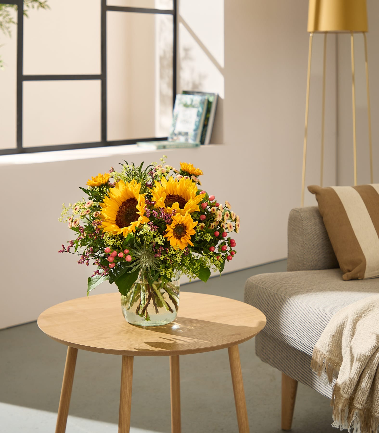 Im Vordergrund steht ein leuchtender Sonnenblumenstrauß auf einem Holztisch, im Hintergrund ein sonnenbeschienenes Fenster und ein gemütliches Sofa. (Beschreibungstext KI-generiert)