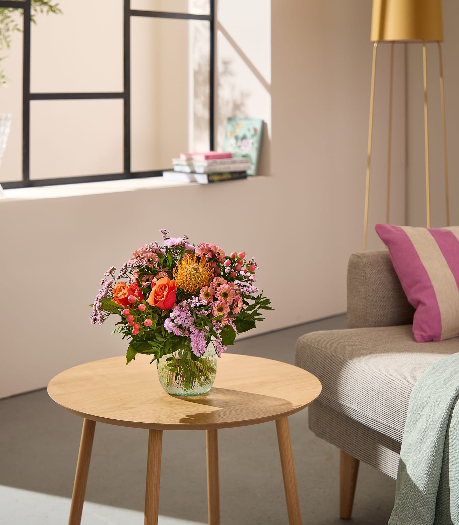 Im Vordergrund steht ein farbenprächtiger Blumenstrauß auf einem Holztisch, im Hintergrund sind ein Sofa und ein Fenster zu sehen. (Beschreibungstext KI-generiert)