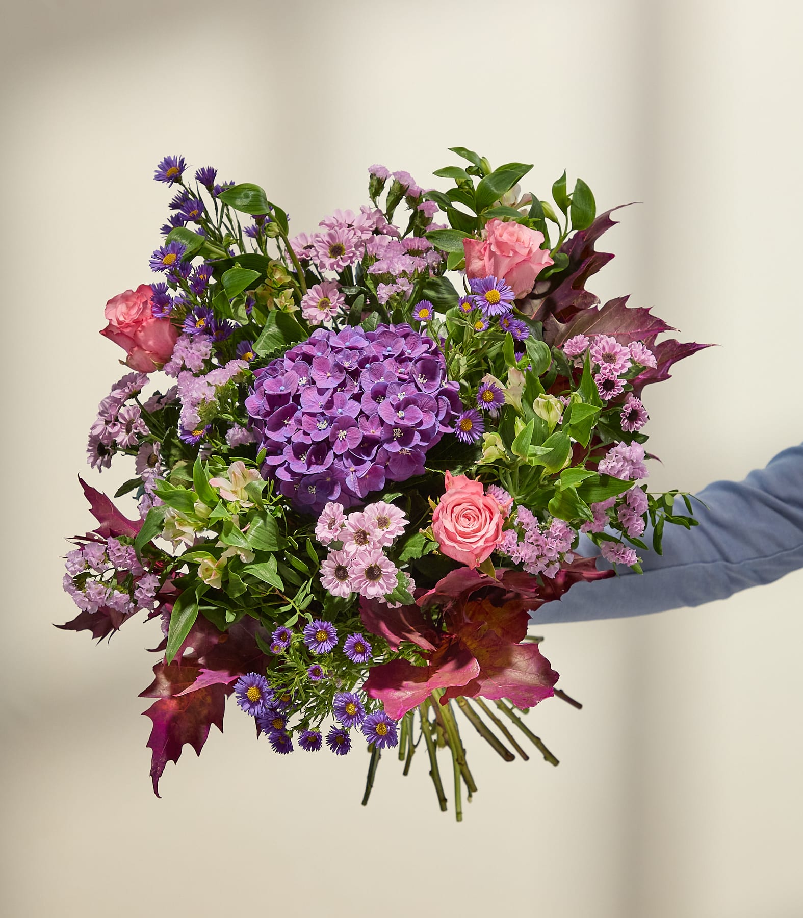 Ein leuchtender Strauß aus lila und rosa Blüten, akzentuiert mit tiefroten Blättern, hebt sich von einem sanft beleuchteten, neutralen Hintergrund ab. (Beschreibungstext KI-generiert)