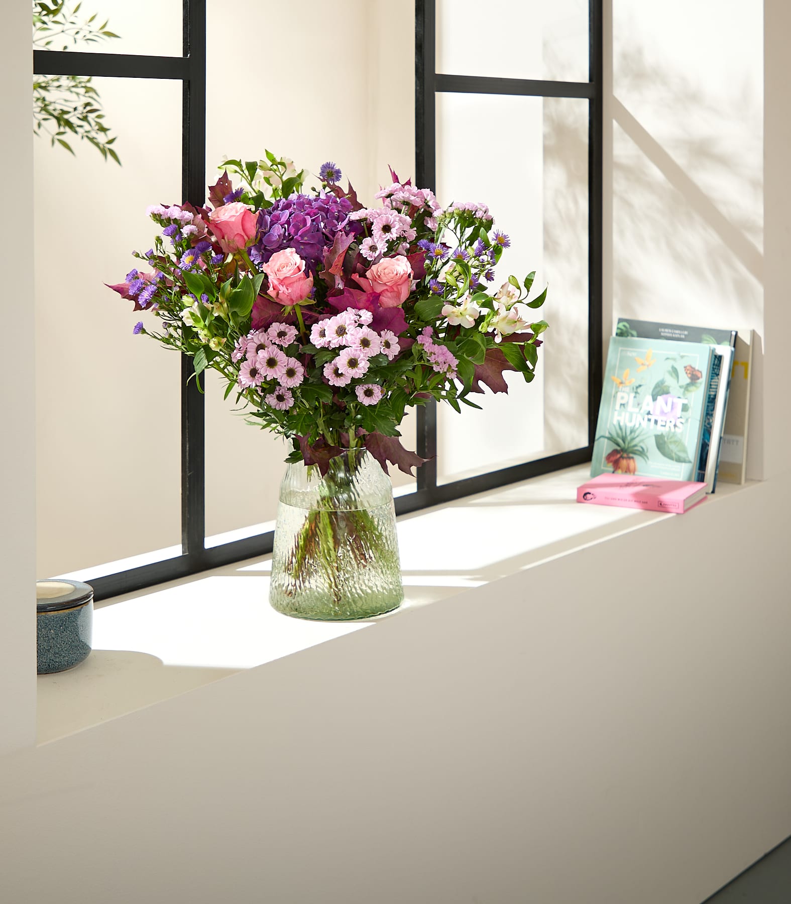 Auf einem Fensterbrett steht ein farbenfroher Strauß rosa und violetter Blumen in einer strukturierten Glasvase neben zwei Büchern; im Hintergrund sind der Fensterrahmen und Grünpflanzen zu sehen. (Beschreibungstext KI-generiert)