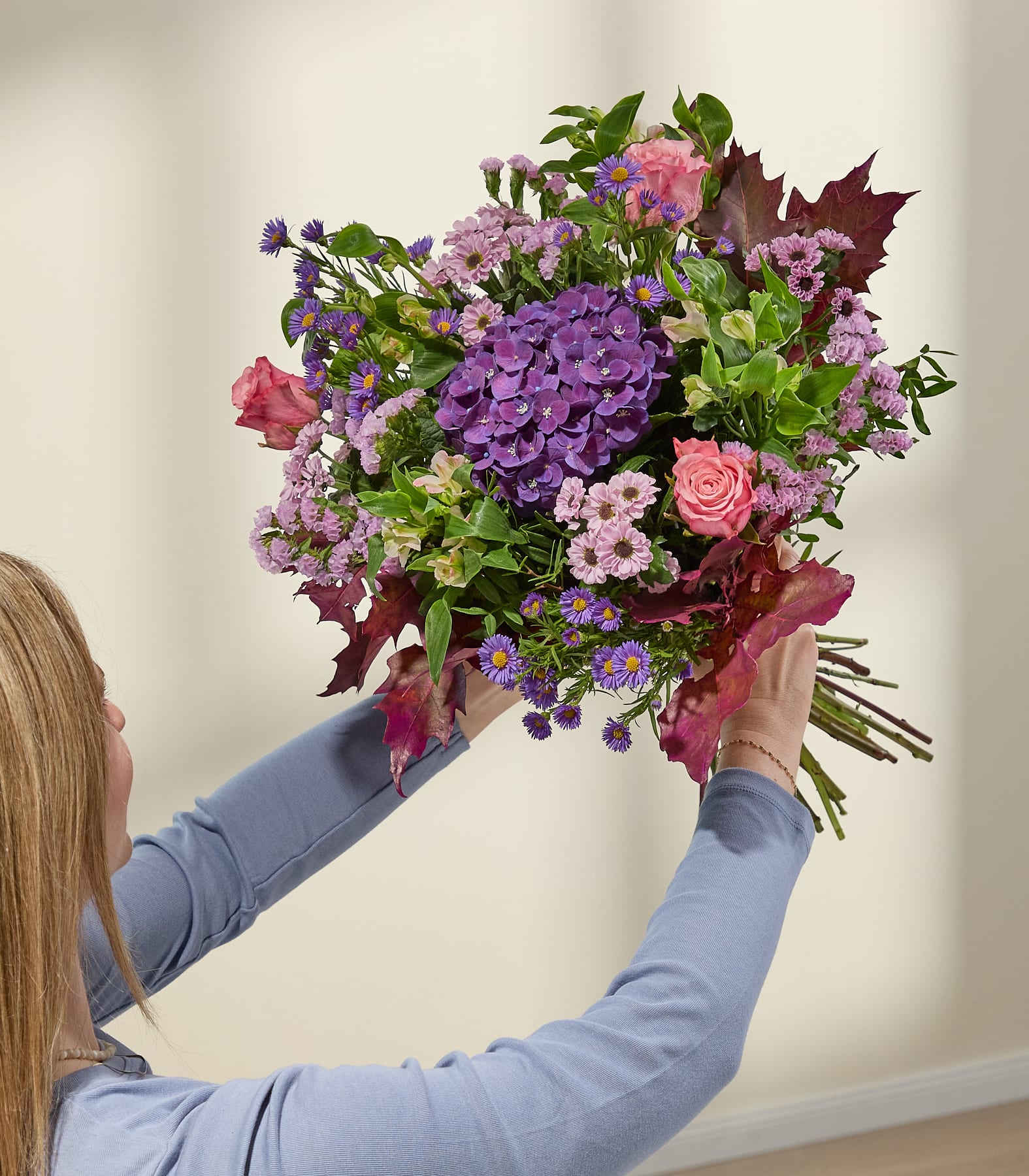 Eine Frau in einem blauen Hemd hält einen leuchtenden Strauß rosa und violetter Blumen, darunter Hortensien und Rosen mit dunkelroten Blättern, vor einer sanft beleuchteten, neutral getönten Wand. (Beschreibungstext KI-generiert)
