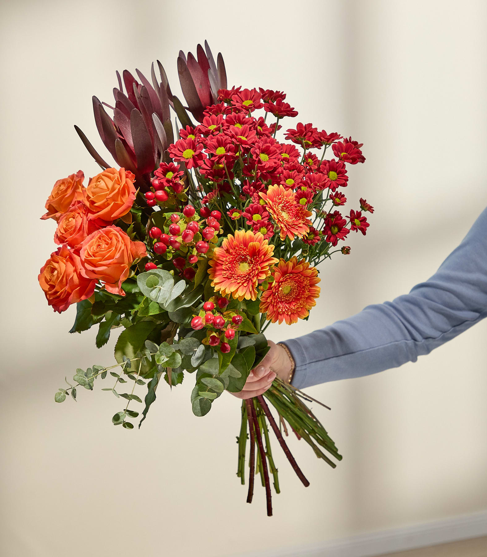 Eine lächelnde Frau in einem weißen Pullover hält vor einem schlichten weißen Hintergrund einen farbenfrohen Strauß roter, orangefarbener und rosafarbener Blumen, darunter Rosen, Gerbera und anderes Grün.