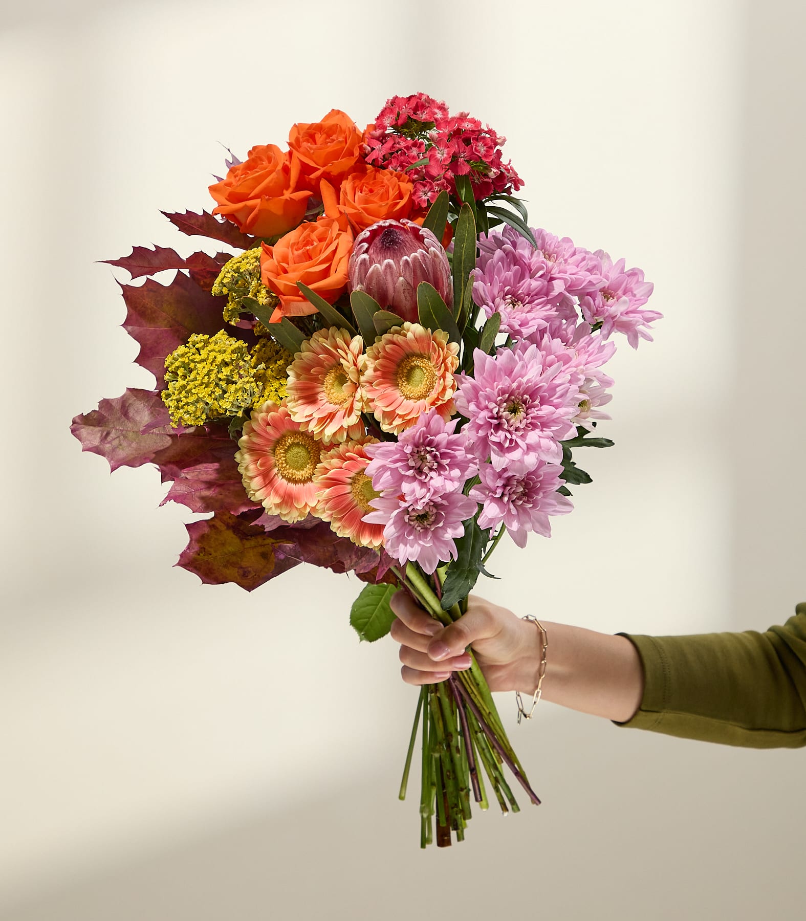 Eine Hand in einem grünen Ärmel hält einen leuchtenden Strauß aus orangefarbenen Rosen, rosa Chrysanthemen und anderen farbenfrohen Blumen vor einem sanft beleuchteten, neutralen Hintergrund. (Beschreibungstext KI-generiert)