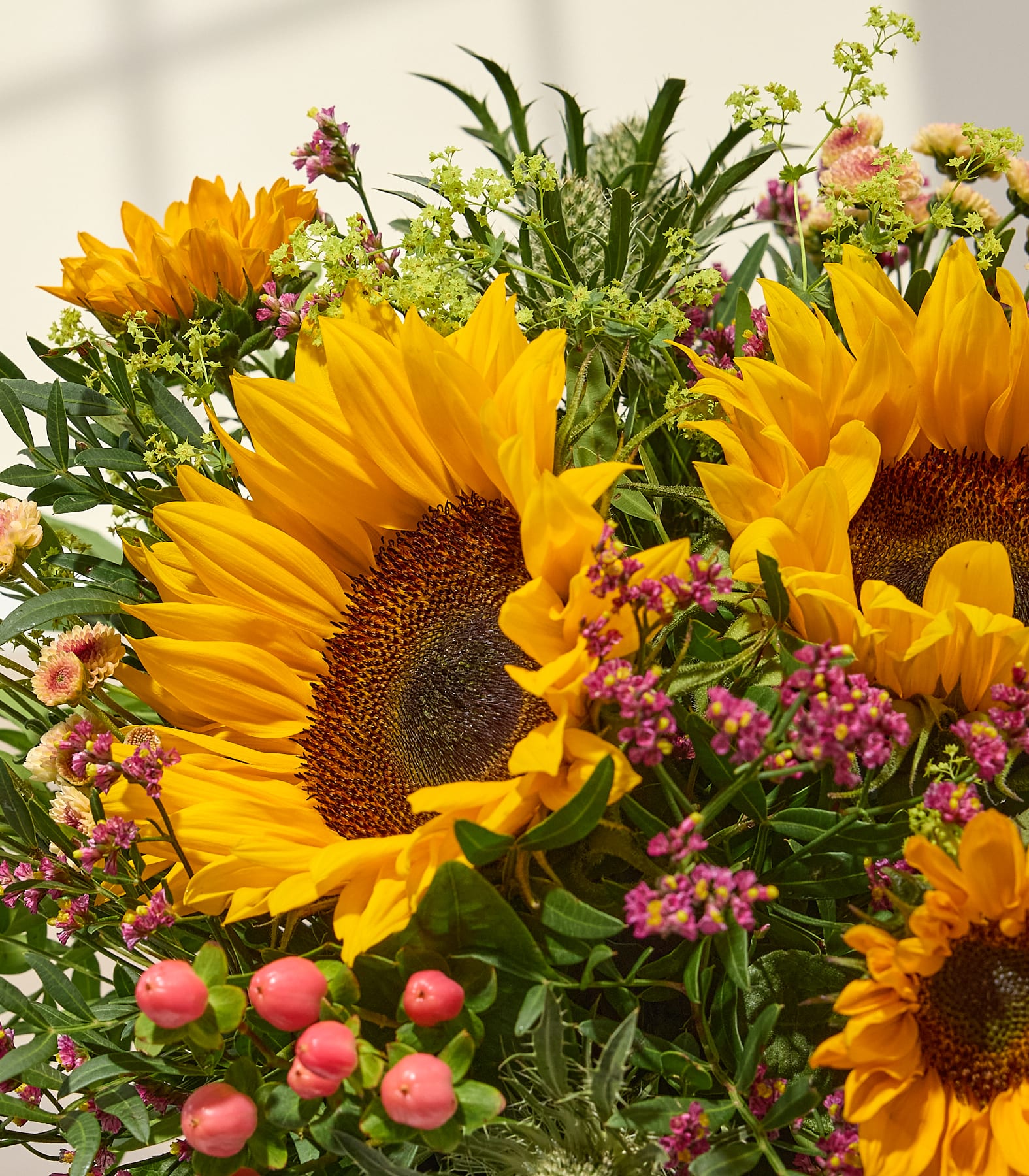 Leuchtend gelbe Sonnenblumen und rosa Beeren füllen den Vordergrund, während ein weicher, schattiger Hintergrund auf eine sanfte Lichtquelle hindeutet. (Beschreibungstext KI-generiert)