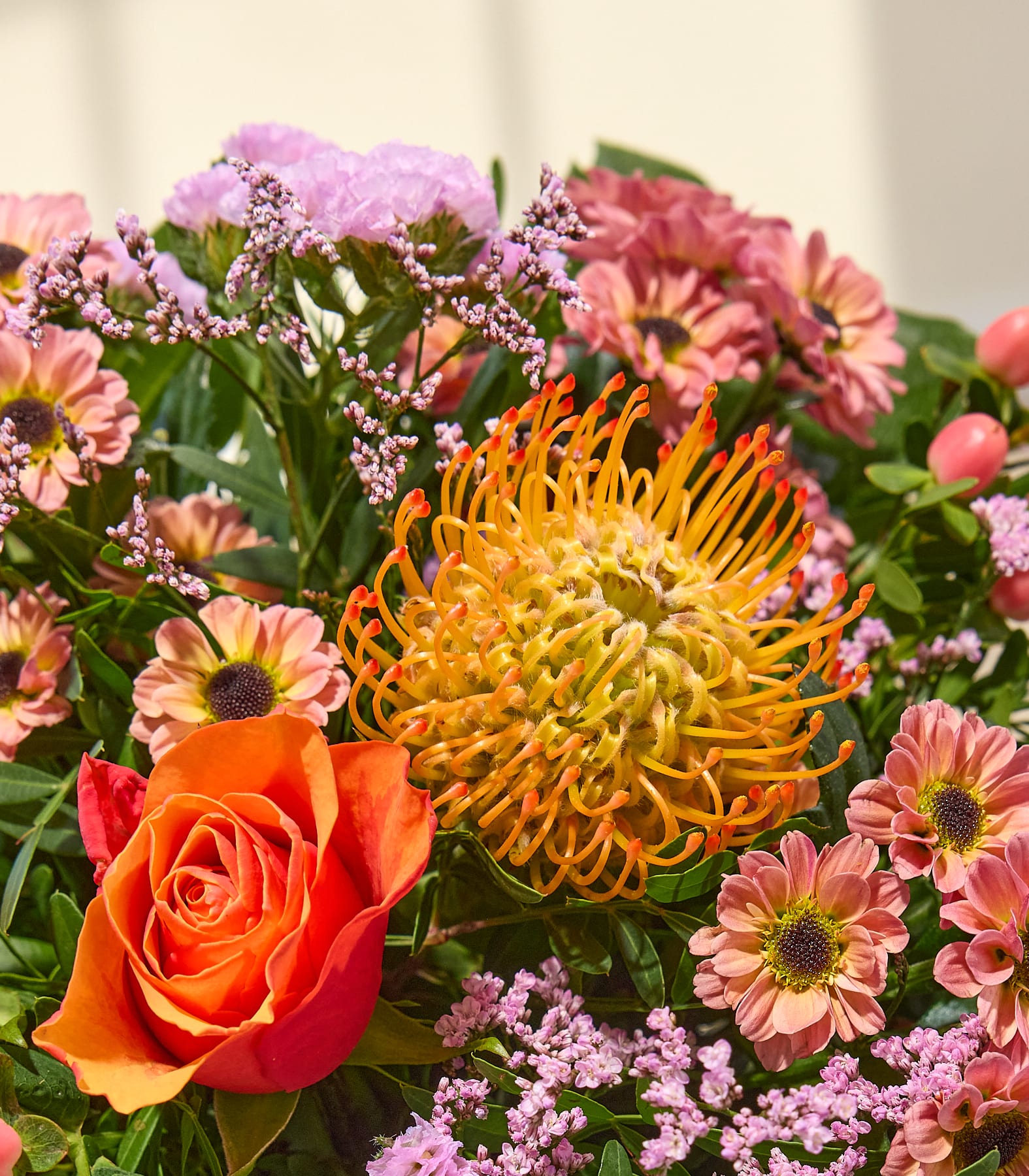 Eine leuchtend orangefarbene Rose und eine stachelige Kissenprotea stechen im Vordergrund eines Straußes aus rosa und violetten Blumen hervor, deren Hintergrund sanft verschwommen ist. (Beschreibungstext KI-generiert)