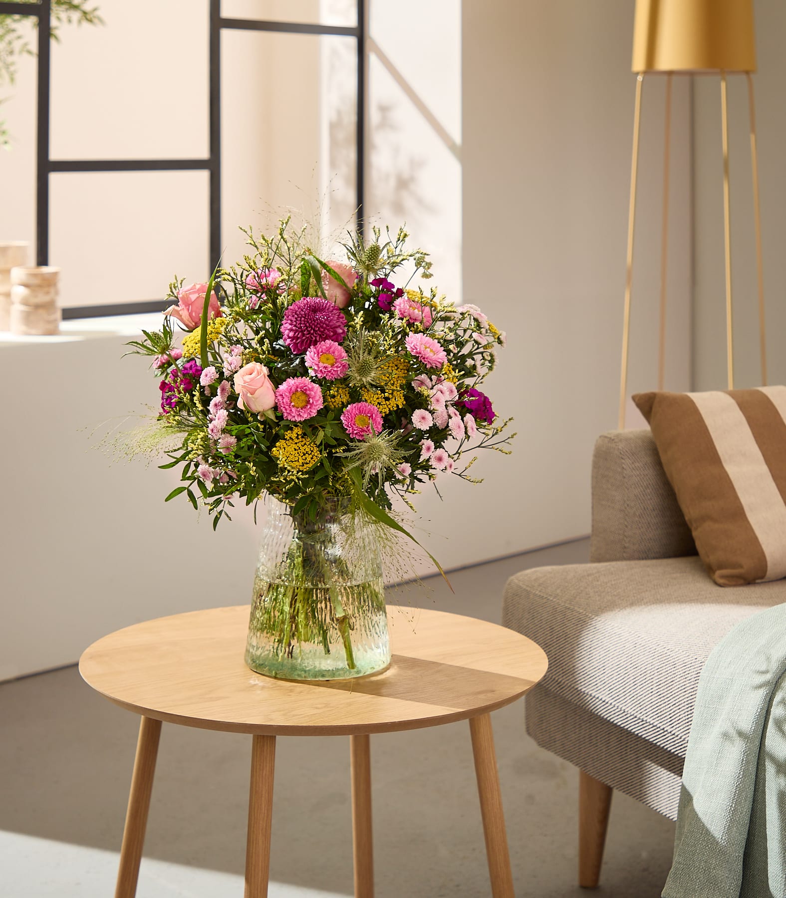 Im Vordergrund steht auf einem hölzernen Beistelltisch ein leuchtender Strauß rosa und gelber Blumen in einer Glasvase; im sanft beleuchteten Hintergrund sind ein Sofa und eine Stehlampe zu sehen. (Beschreibungstext KI-generiert)