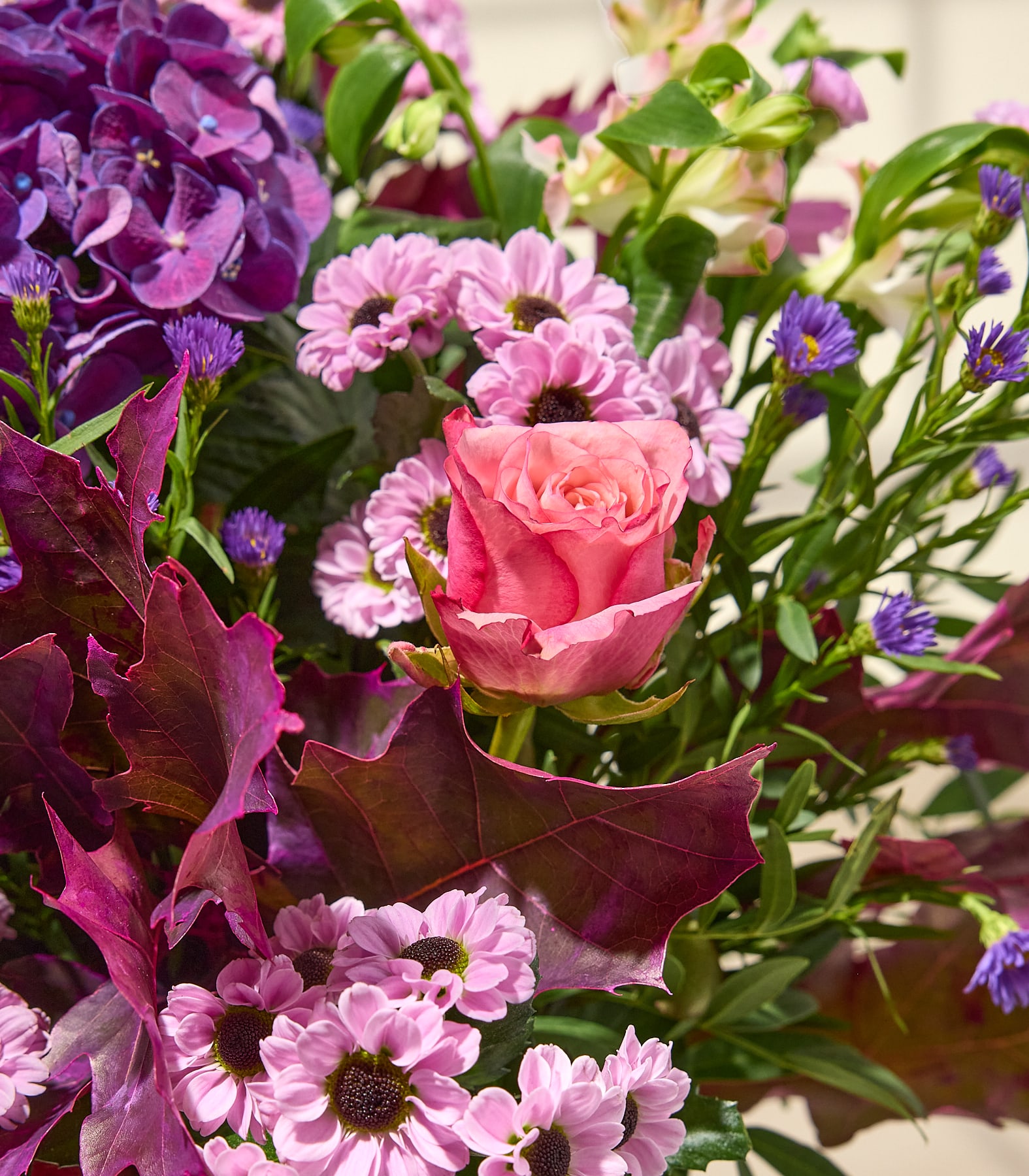 Eine Nahaufnahme eines farbenprächtigen Blumenstraußes zeigt im Vordergrund eine rosa Rose und eine violette Hortensie, während im Hintergrund zarte rosa und violette Wildblumen und grünes Laubwerk zu sehen sind. (Beschreibungstext KI-generiert)