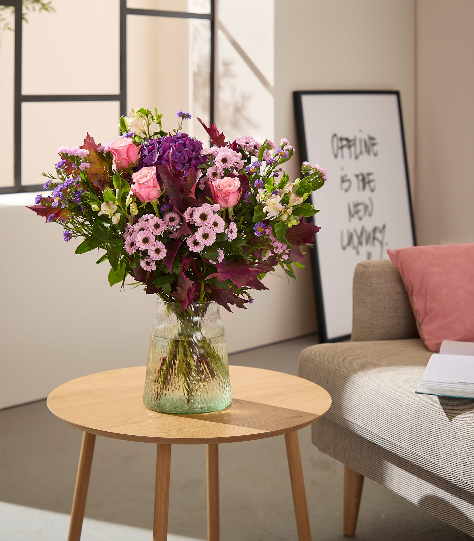 Ein farbenfroher Strauß rosa und violetter Blumen in einer strukturierten Glasvase steht auf einem hölzernen Beistelltisch vor einem Sofa mit einem gerahmten Druck und einem Fenster. (Beschreibungstext KI-generiert)