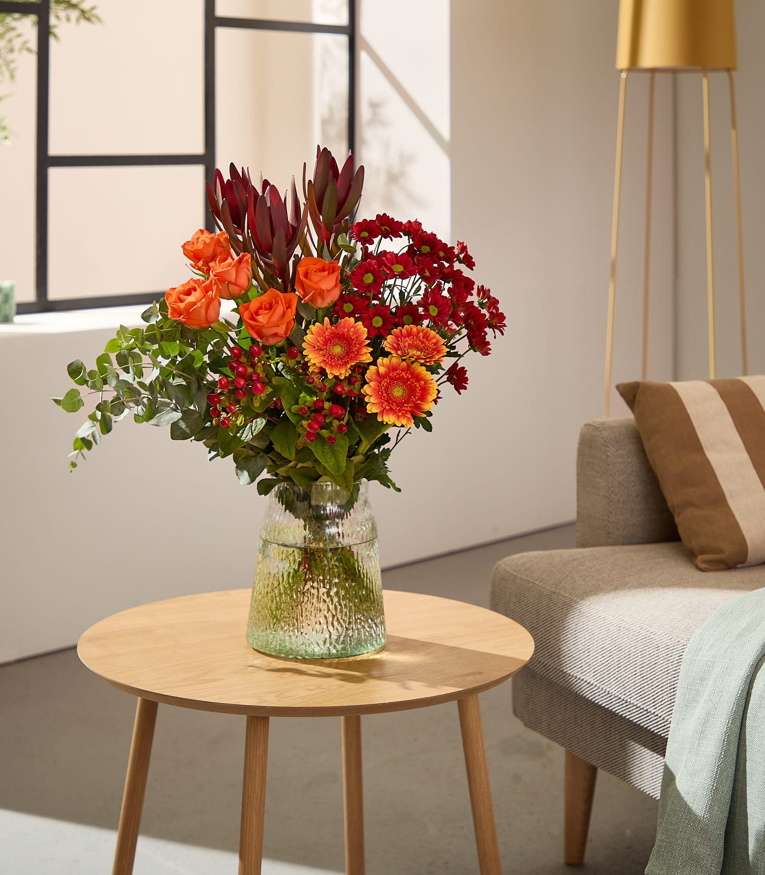 Eine lächelnde Frau in einem weißen Pullover hält vor einem schlichten Hintergrund einen farbenfrohen Strauß roter, orangefarbener und rosafarbener Blumen.