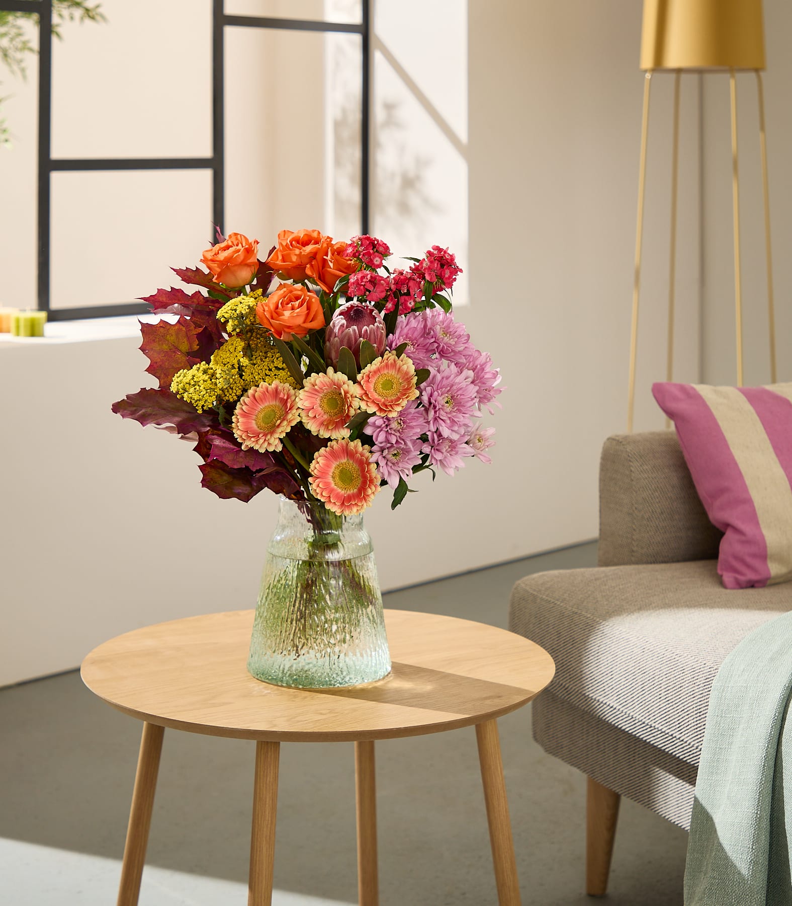 Im Vordergrund steht ein farbenprächtiger Strauß Herbstblumen in einer Glasvase auf einem runden Holztisch; im sanft beleuchteten Hintergrund sind ein Sofa und eine Stehlampe zu sehen. (Beschreibungstext KI-generiert)