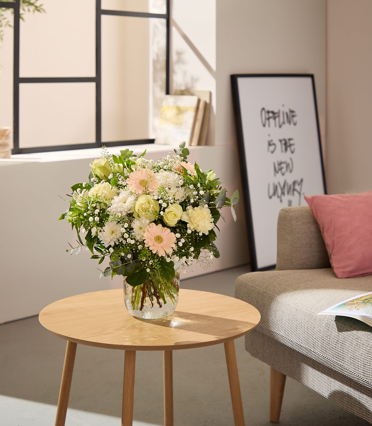 Ein farbenfroher Strauß weißer und rosa Blumen steht in einer Glasvase auf einem hölzernen Beistelltisch, im sanft beleuchteten Hintergrund sind ein gerahmtes Zitat und eine Couch zu sehen.
