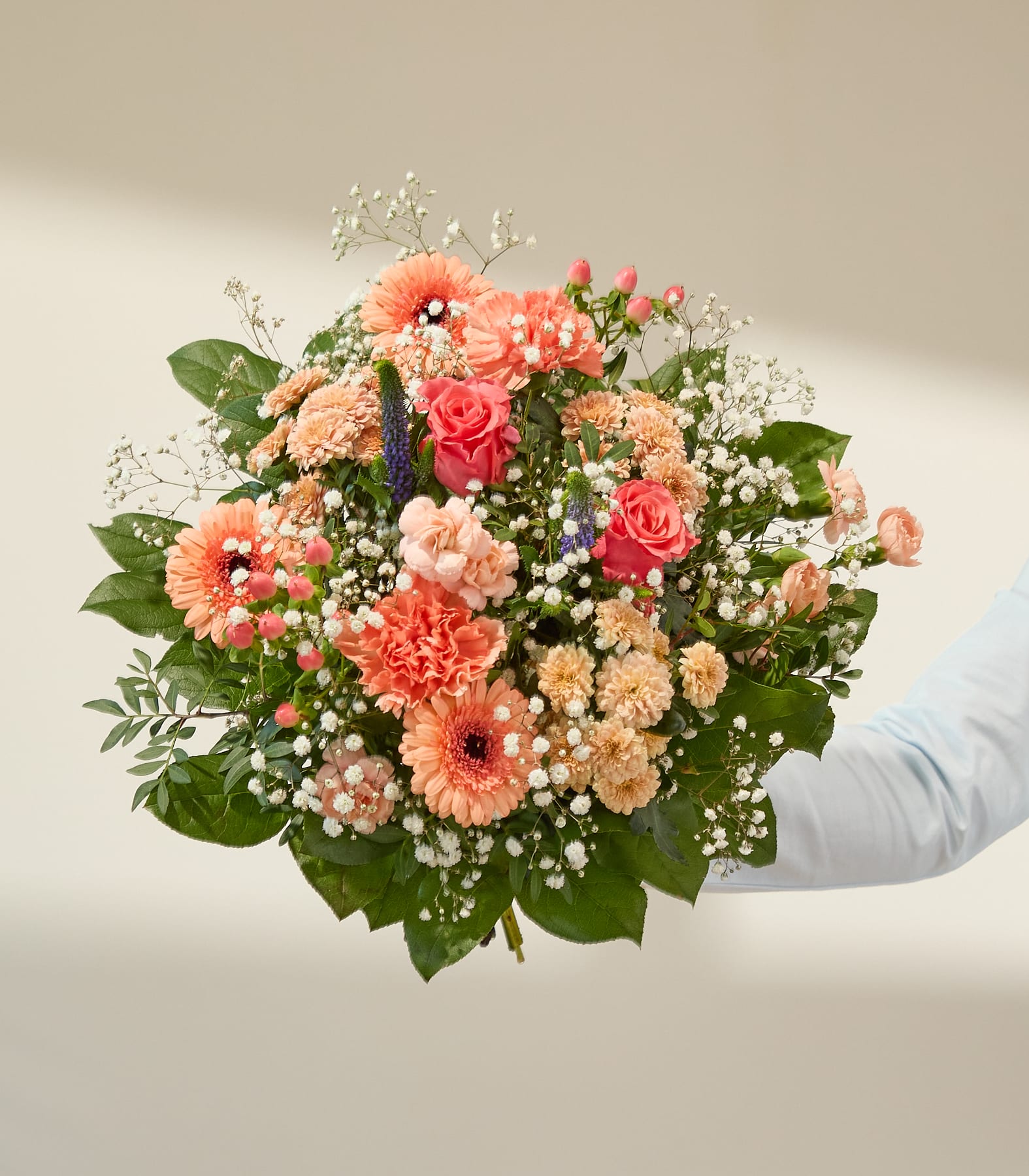 Ein lebhafter Blumenstrauß in Pfirsich- und Rosatönen, akzentuiert mit Schleierkraut und Grünzeug, hebt sich von einem sanft beleuchteten, neutralen Hintergrund ab.