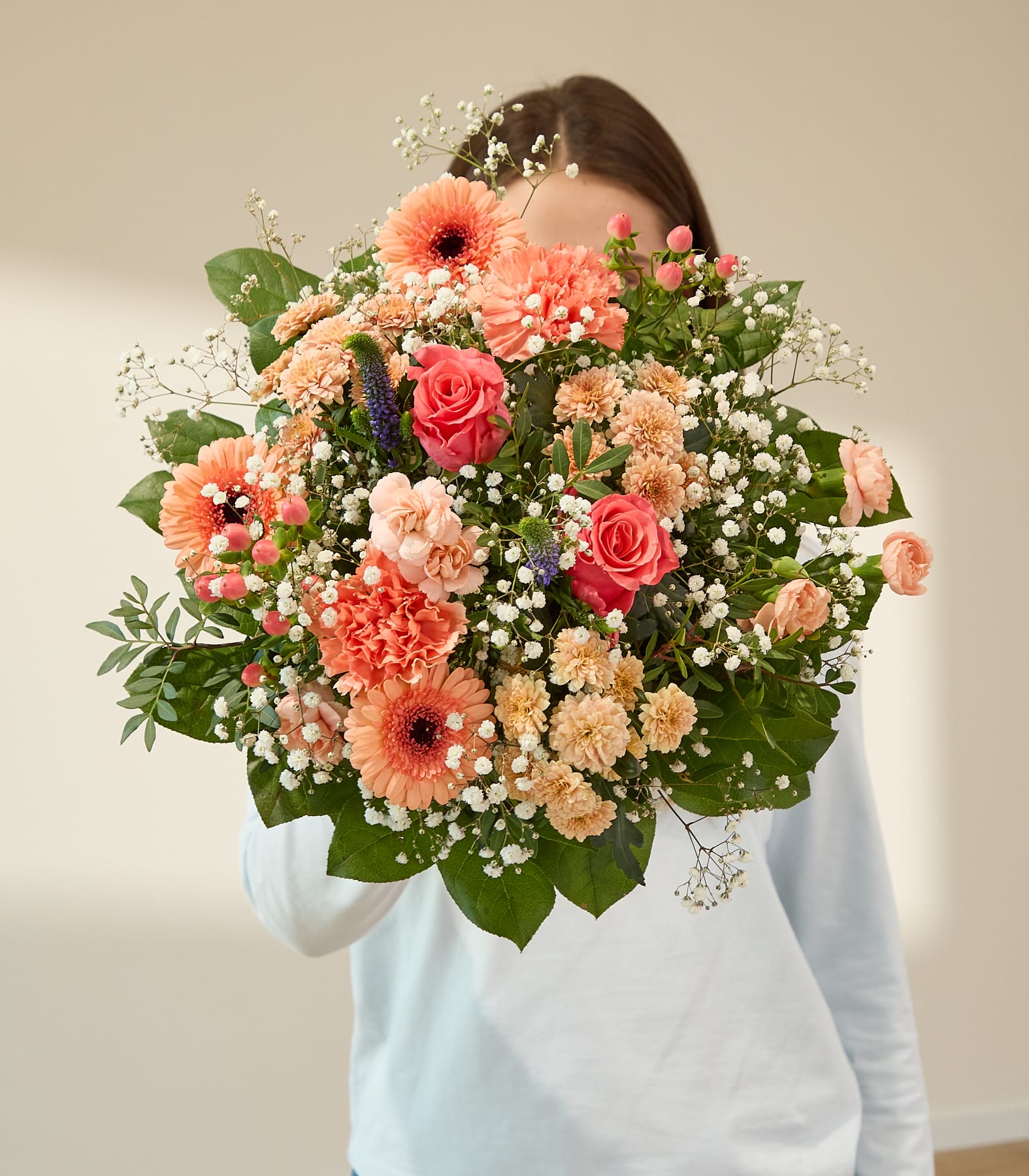 Eine Person in einem weißen Pullover hält einen großen Strauß rosa und orangefarbener Blumen; im Hintergrund ist eine schlichte, hellfarbige Wand zu sehen.