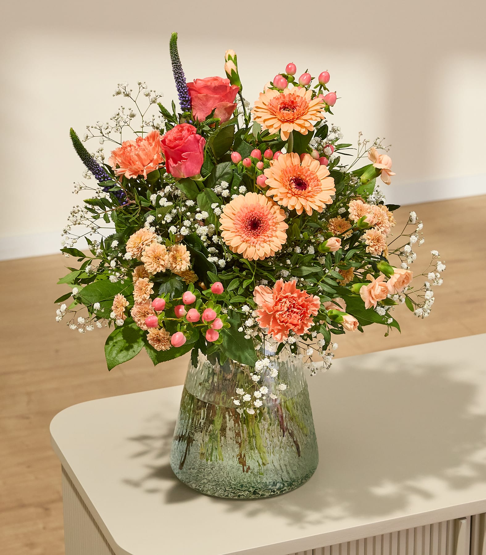 Ein farbenfroher Blumenstrauß aus verschiedenen Blumen, darunter Gerbera, Rosen und Nelken, der von einer Person in den Händen gehalten wird, vor einem schlichten weißen Hintergrund. (Beschreibungstext KI-generiert)