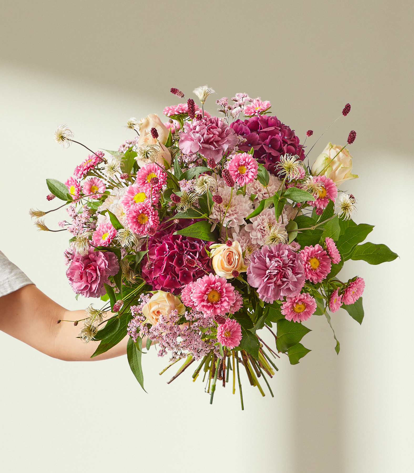 Eine Hand hält einen leuchtenden Strauß rosa und violetter Blumen vor einem sanft beleuchteten, neutralen Hintergrund. (Beschreibungstext KI-generiert)