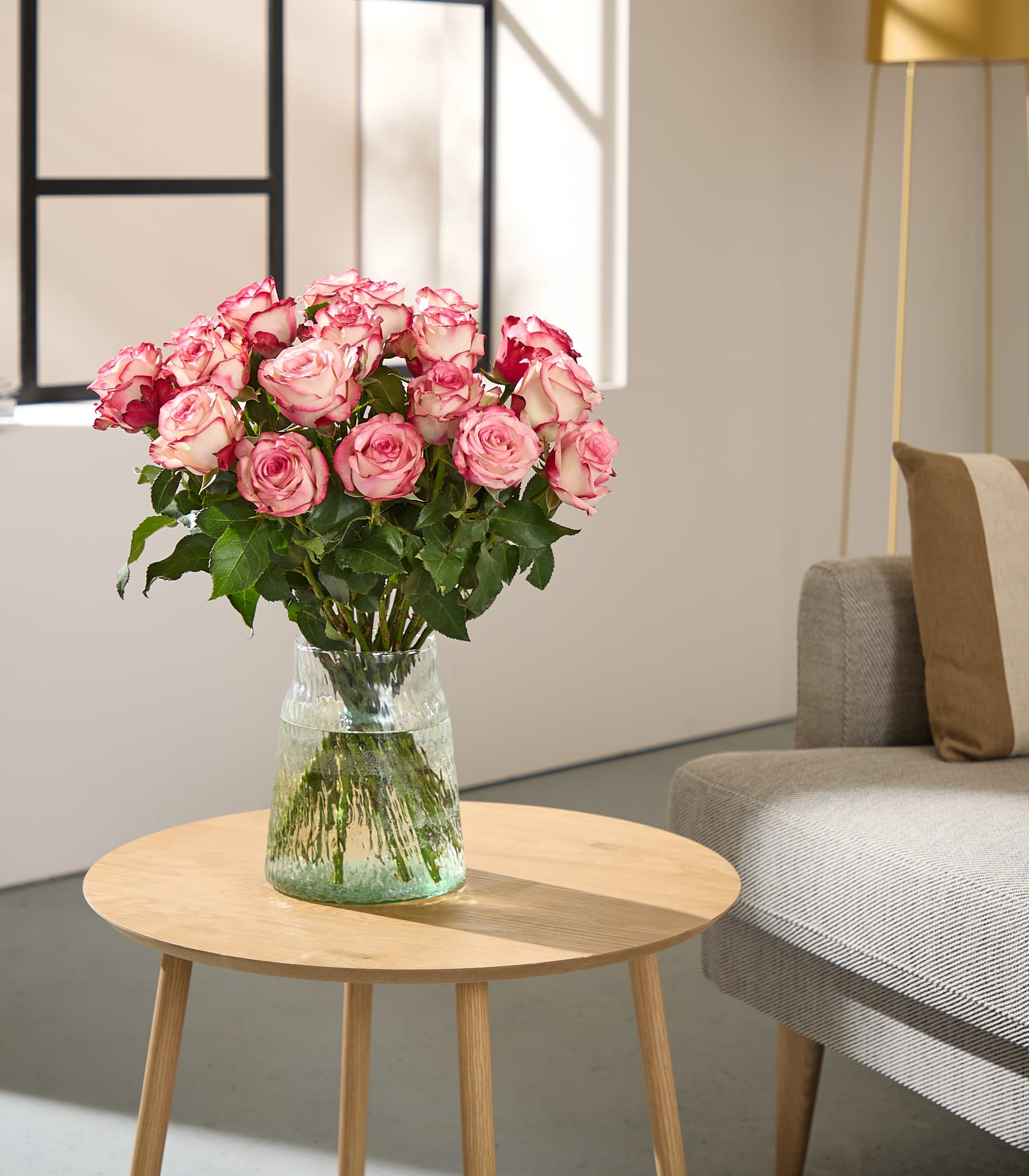 Auf einem hölzernen Beistelltisch in einem gemütlichen Wohnzimmer steht ein Strauß leuchtend pinkfarbener Rosen; ein graues Sofa und ein großes Fenster sorgen für natürliches Licht.